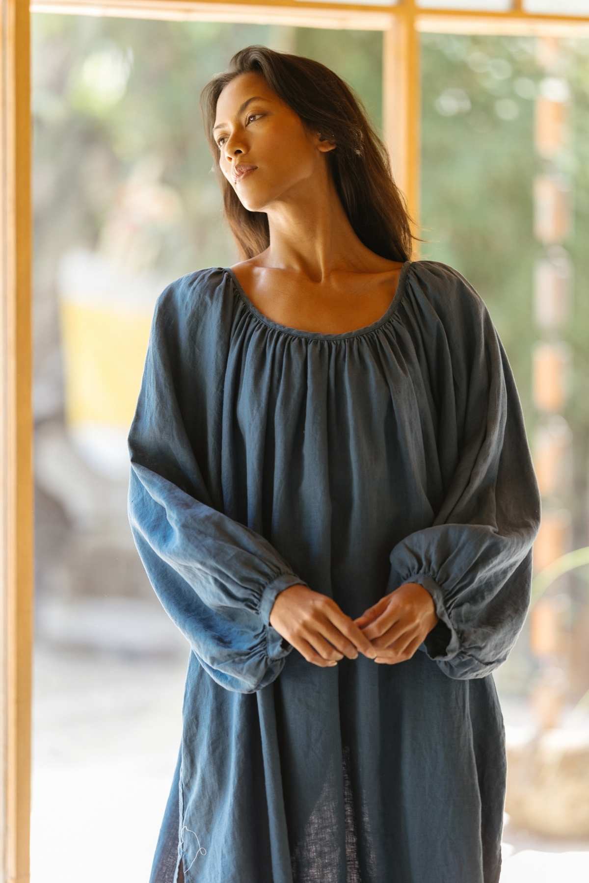 A woman with long dark hair stands indoors near a large window, looking thoughtfully into the distance. She is wearing a loose, dark blue Alchemy Gown made of crinkle linen cotton from Myrah Penaloza, embodying the essence of a modern mystic. The background is softly lit with natural light.