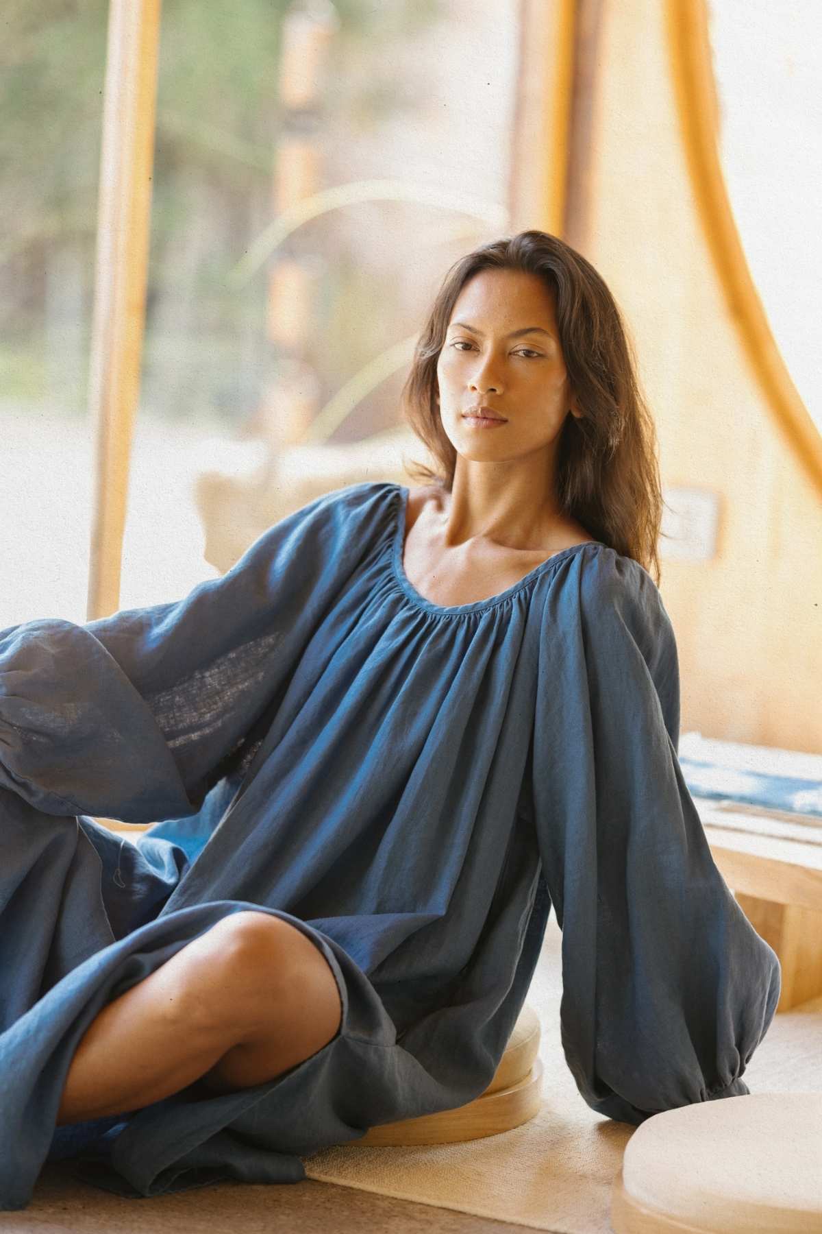 A woman with long dark hair sits on the floor beside a wooden structure. She is wearing Myrah Penaloza's Alchemy Gown (Crinkle Linen Cotton) (PR), which features a loose, long-sleeved blue design and looks relaxed. Sunlight filters through the windows in the background, creating a warm, serene ambiance.