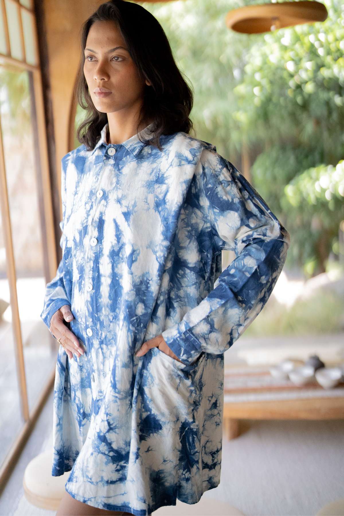 A person stands indoors wearing a unique piece—a Tender Jacket by Myrah Penaloza, handcrafted with hand-dyed indigo. They have long dark hair, hands resting in their pockets as they gaze thoughtfully into the distance. The background showcases a window framing lush greenery and a wooden table.