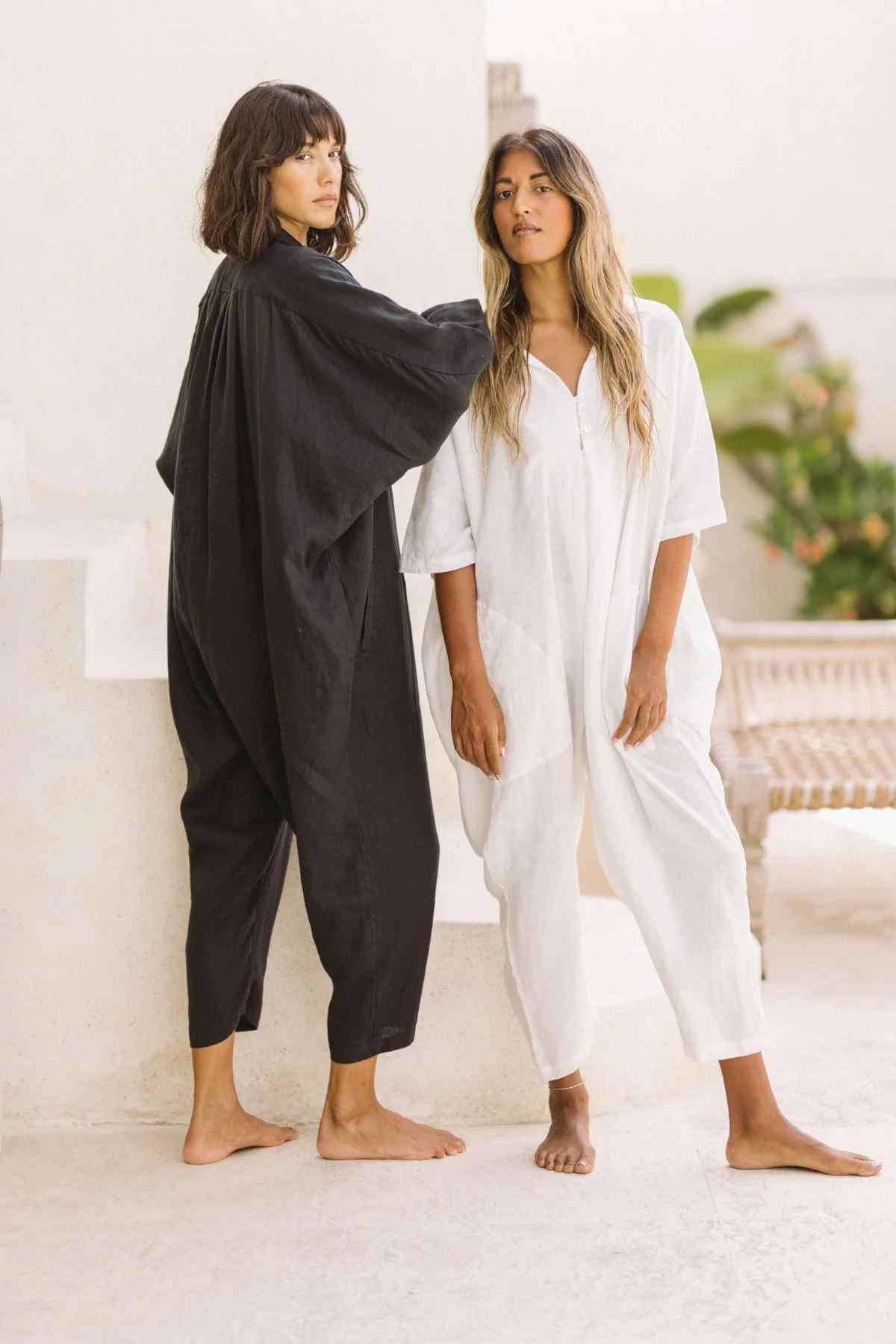 Two women are standing side by side in a minimalist, light-filled space. One woman wears a black, loose-fitting Linen Sat Torri Playsuit with wide dolman sleeves from Myrah Penaloza, while the other sports a similar playsuit in white. They are both barefoot and gazing at the camera with neutral expressions, embodying a retro 1980s inspired design.