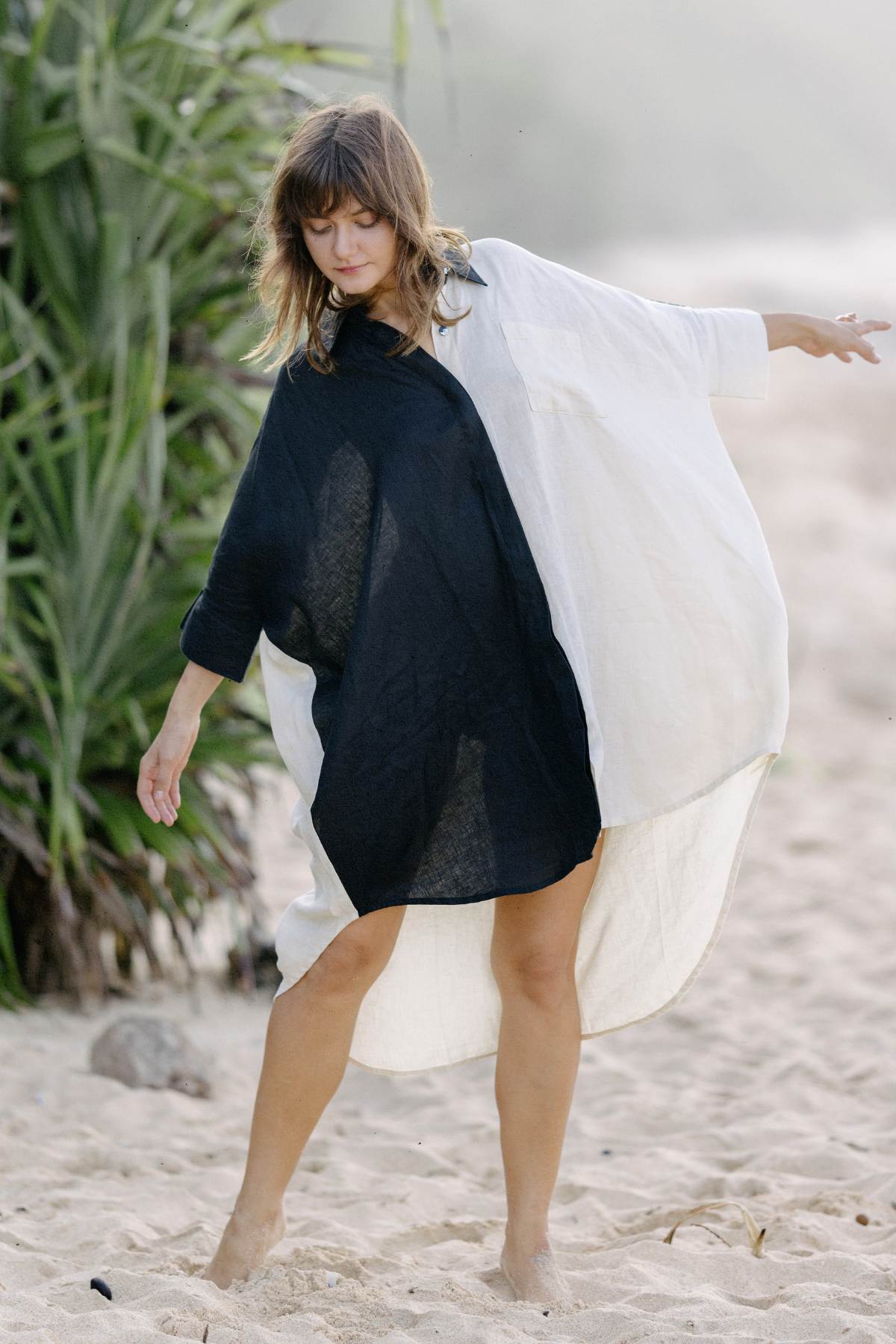 A woman with shoulder-length hair, wearing the Myrah Penaloza Linen Suka Button Down Long Top in black and white, is standing barefoot on a sandy beach. She has one arm outstretched and appears to be moving gracefully. Green foliage is visible in the background.