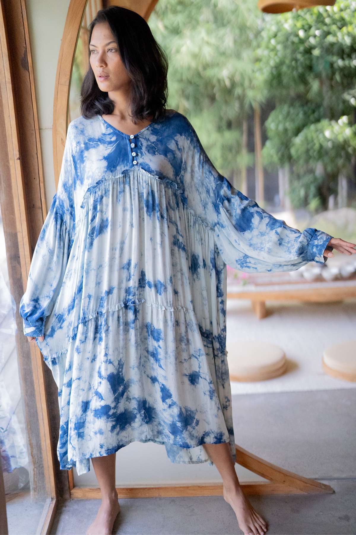 A person wearing the Myrah Penaloza Indigo Eva Gown, made from 100% silk with an oversized fit, stands barefoot by a large window framed with wood. Their shoulder-length dark hair gently cascades as they gaze thoughtfully outside towards the serene green outdoor backdrop.