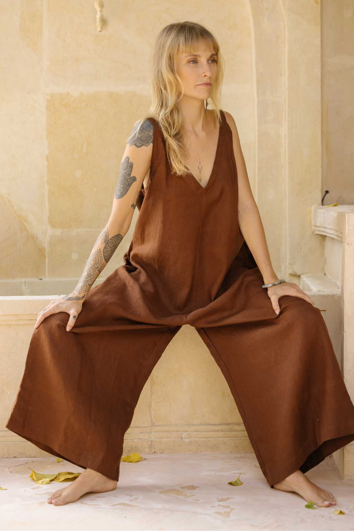 A person with long blonde hair and tattoos on their right arm is sitting on a ledge in front of a beige-colored wall. They are wearing the luxuriously soft Myrah Penaloza Kundalini Linen Playsuit, which features wide-leg bottoms and is reversible. They are barefoot, gazing thoughtfully into the distance. Fallen leaves are scattered on the floor.