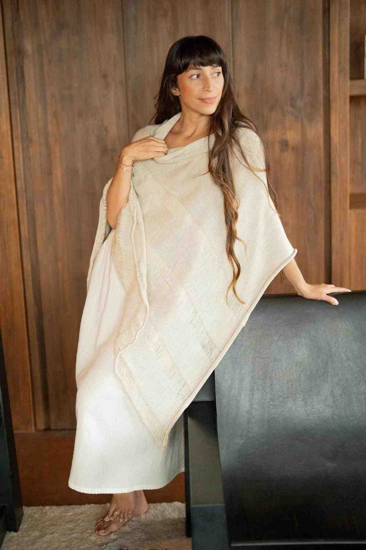 A woman with long hair sits on the arm of a dark chair, wrapped in an effortlessly chic, cream-colored Frida Poncho by Myrah Penaloza. She is gazing to her left. The setting appears to be indoors with wooden-paneled walls in the background, making her poncho look like a timeless wardrobe staple.