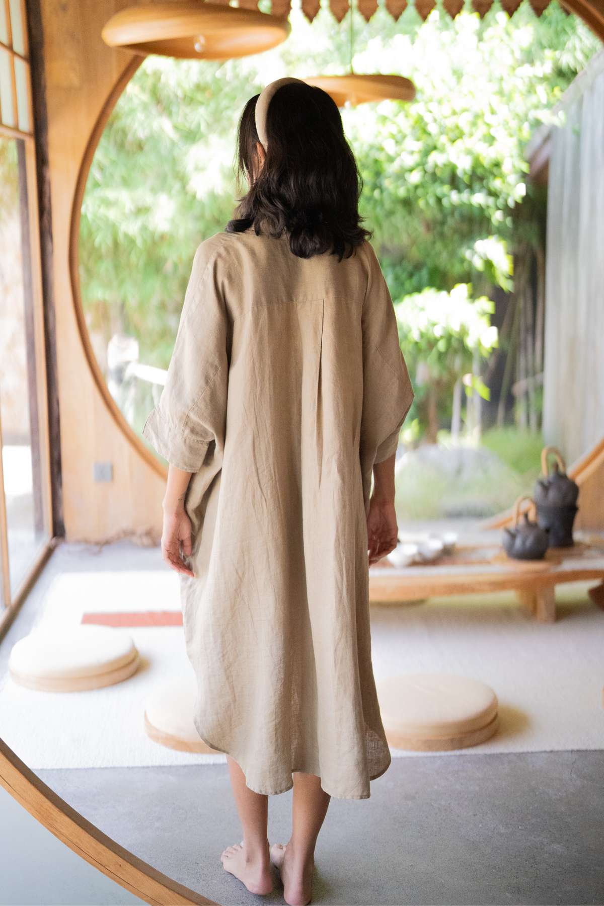 A person with long dark hair, dressed in a Linen Suka Button Down Long Top - BF by Myrah Penaloza made of sustainable fabric, stands barefoot inside a minimalist, circular wooden doorway. The room is softly lit, with green plants outside and a tea set visible in the background.