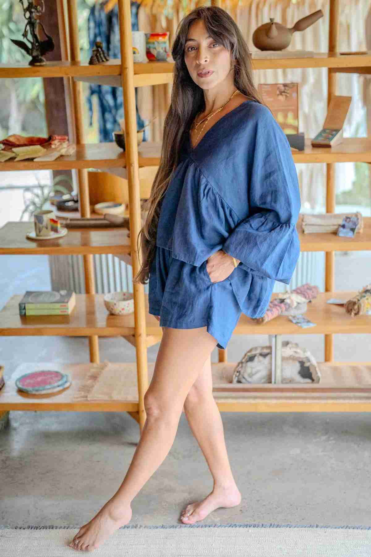 A woman with long hair stands barefoot on a rug in a well-lit room, wearing an Eva Linen Top from Myrah Penaloza with flowering sleeves and matching shorts. Behind her is a wooden shelf filled with various items, including books, pottery, and decor. She has one hand in her pocket and is smiling slightly.