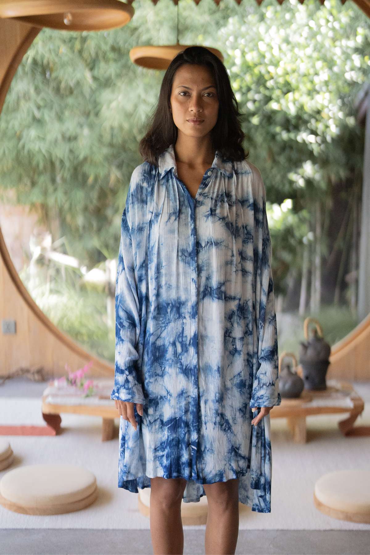 A person wearing the Indigo Dreams Kundalini Gown Short by Myrah Penaloza stands indoors, with circular wooden decor and lush greenery visible in the background.