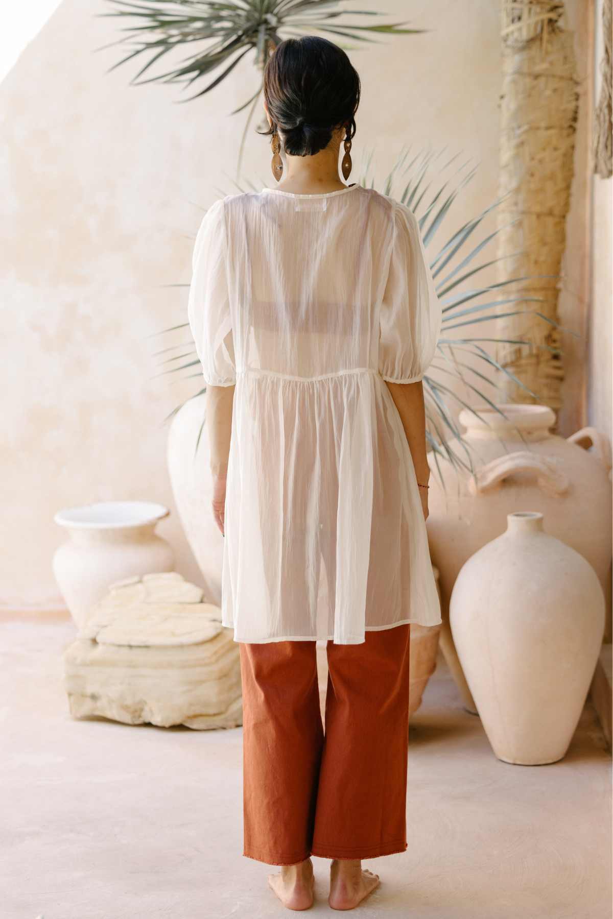 A woman stands indoors with her back to the camera, showcasing the Dynasty Sheer Silk Dress by Myrah Penaloza and rust-colored pants. Her hair is tied up, and she accessorizes with large earrings. The background features large white pottery and greenery.