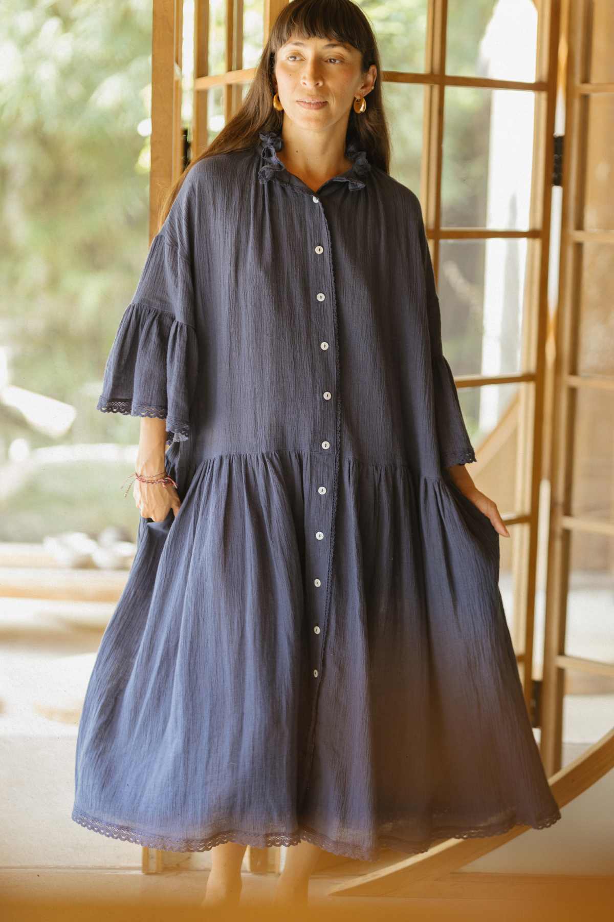 A woman stands indoors wearing the Kyoto Duster, a made-to-order piece by Myrah Penaloza. The duster features a loose, flowing design in navy blue with a button-down front and ruffled sleeves, evoking the elegance of ceremonial wear. She holds the sides of the attire as light streams through large windows in the background.