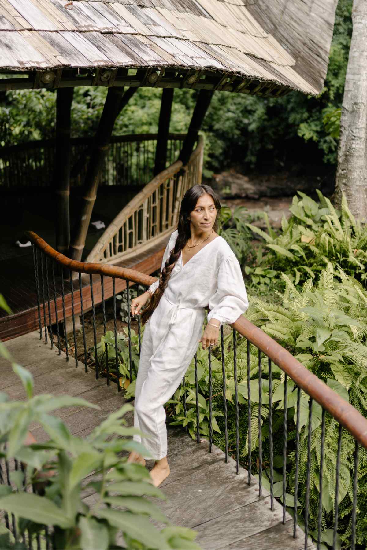 A person with long hair sits cross-legged on a rug indoors, wearing a white 100% Linen Long Sleeve Dora Playsuit by Myrah Penaloza, featuring an adjustable waistline. They have their eyes closed and hands together in front of their face as if meditating or practicing yoga. Sunlight filters through the bamboo structure in the background.