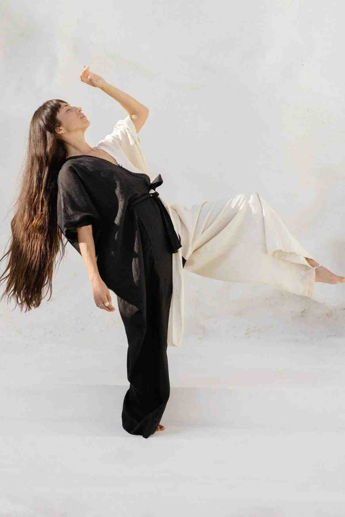 A person with long, flowing hair is captured mid-motion, wearing a black and white French Linen Yin Yang River Playsuit from Myrah Penaloza. The individual's left leg is raised, and their right arm is bent upward against a neutral, light background. The scene conveys a sense of balance and fluidity.