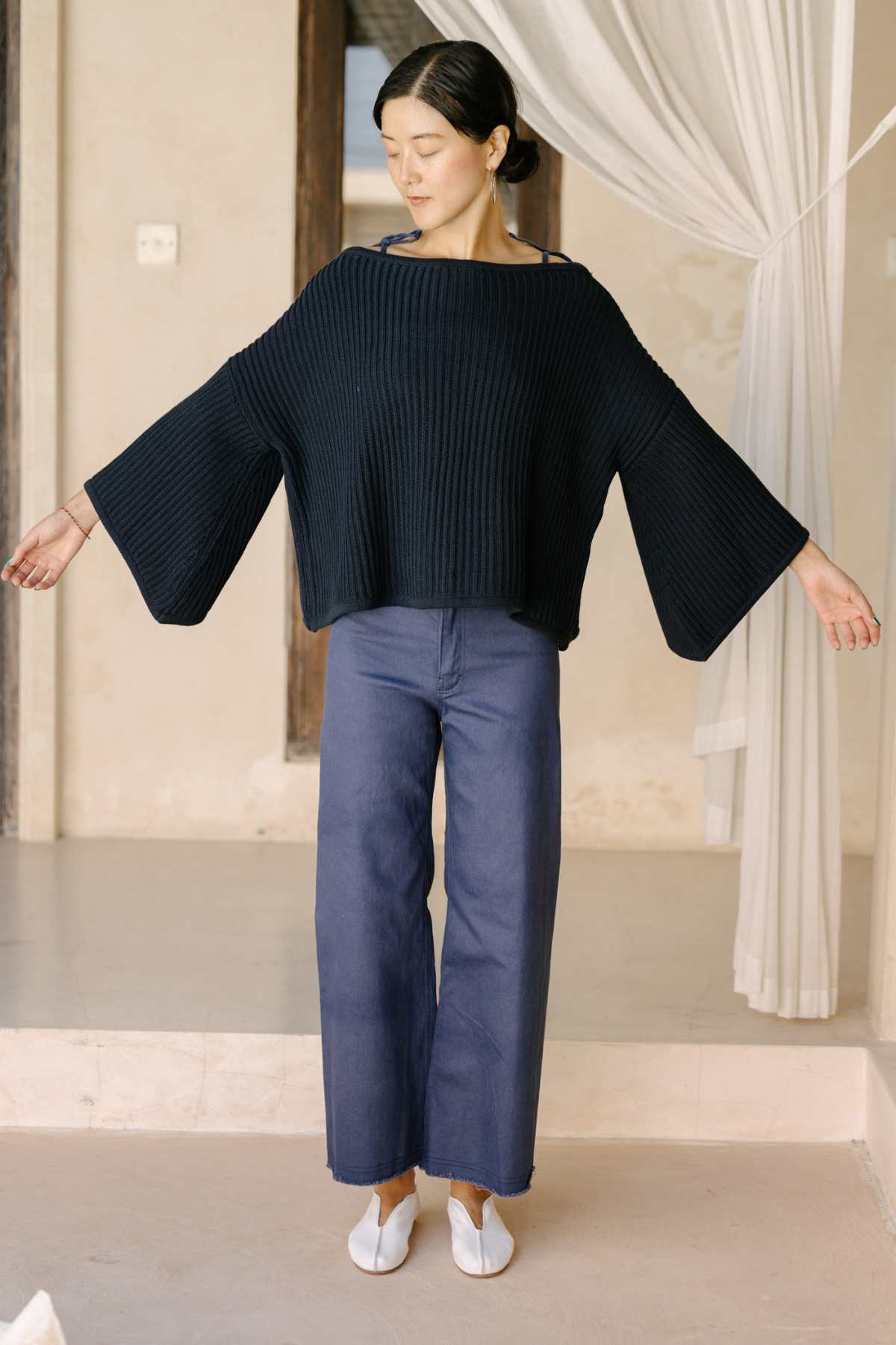 A person stands indoors with arms outstretched, wearing a black Spirit Top Sweater Knit by Myrah Penaloza, featuring ribbed textures and wide sleeves, paired with blue wide-leg pants and white shoes. The background showcases a neutral-toned interior with a window adorned with sheer curtains.