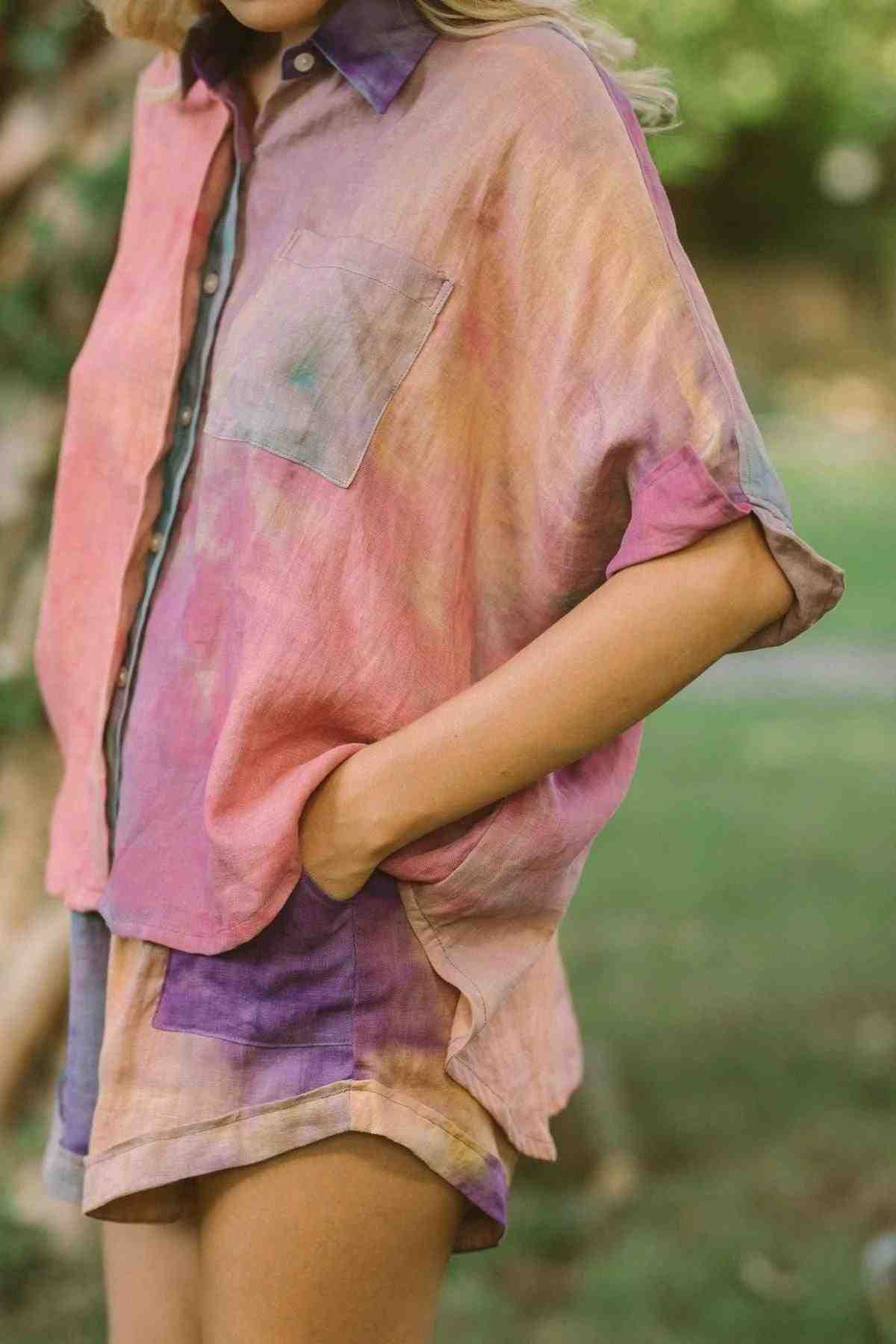 A person wearing the Suka Button Down Top from Myrah Penaloza's Rainbeau Collection, made from 100% linen and boasting a colorful, tie-dye design with matching shorts. The ethically sourced clothing has a loose-fitting, worn-in look against a blurred green outdoor background.