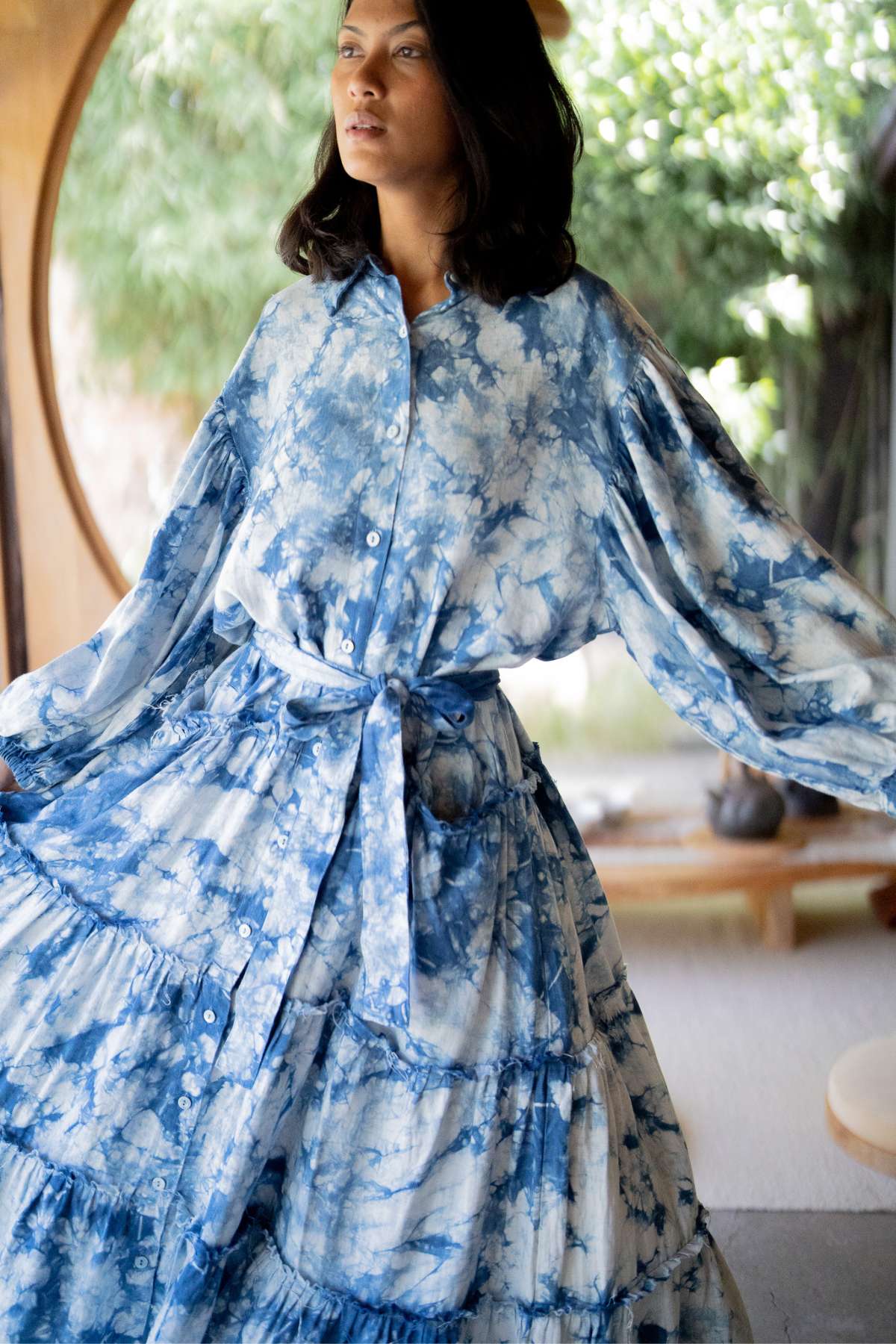 A woman stands indoors wearing the La Dama Long Sleeves Ruffles gown by Myrah Penaloza, her hands gracefully lifting the fabric of the long, flowing indigo dress with a belt. Her gaze drifts to the side, framed by a round window displaying lush greenery.