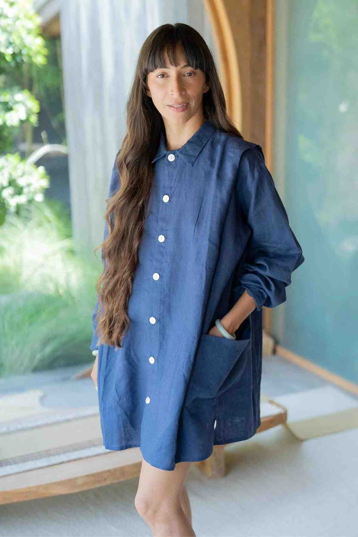 A woman with long, wavy brown hair stands indoors wearing a deep royal blue Tender Jacket from Myrah Penaloza. The 100% linen jacket features large pockets and buttons up the front. She has a relaxed expression and stands with one hand in her pocket. A background of greenery and a glass window is partially visible, enhancing the eclectic piece she dons.