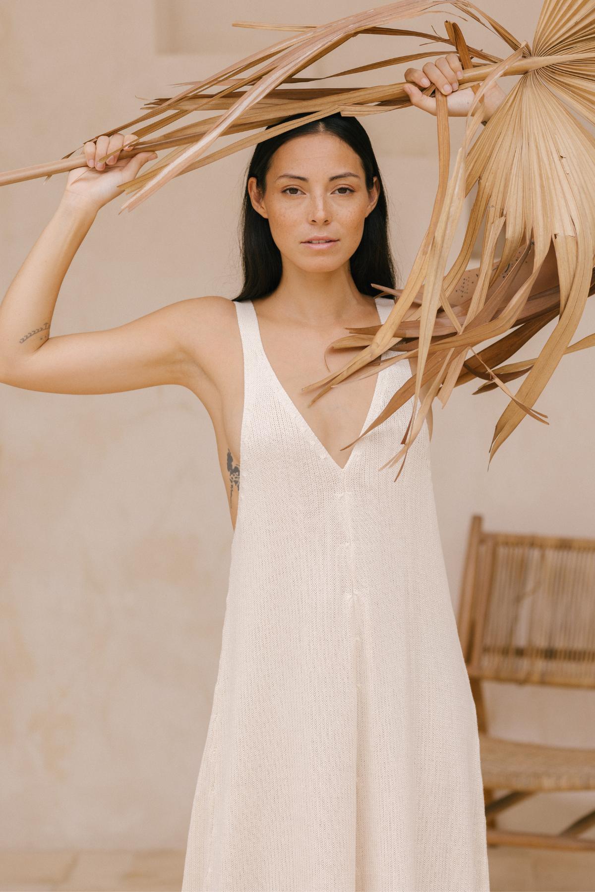 Wearing the Kundalini Playsuit by Myrah Penaloza, a person in a white sleeveless outfit artfully displays dried palm leaves above their head, radiating effortless style and versatility. The setting, featuring a light-colored wall and a wooden chair, encapsulates a natural and minimalist aesthetic.