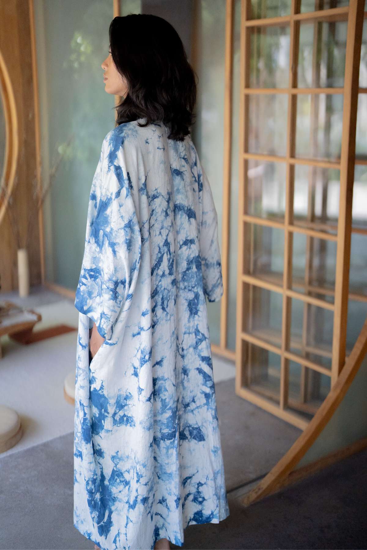 A person with shoulder-length dark hair is seen indoors, standing near a large wooden door with glass panels while wearing Myrah Penaloza's The Boyfriend Dress, hand tie-dyed in indigo. The room features soft natural lighting and minimalistic decor, and the person is facing away from the camera.