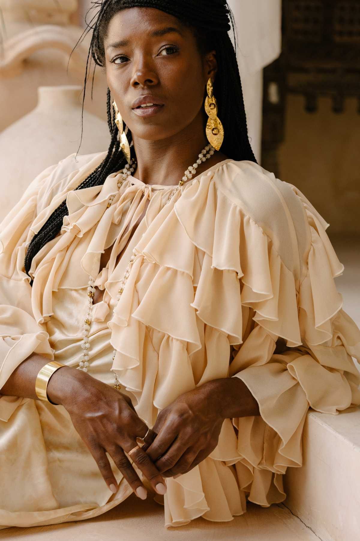 A woman with braided hair wearing statement gold earrings and a layered, ruffled beige blouse is seated in a poised manner. She exudes confidence and sophistication, complemented by a serene background with neutral tones. Her ensemble, Anusara Bloom - Made To Order by Myrah Penaloza, adds an elegant touch reminiscent of Nature-Inspired Couture.