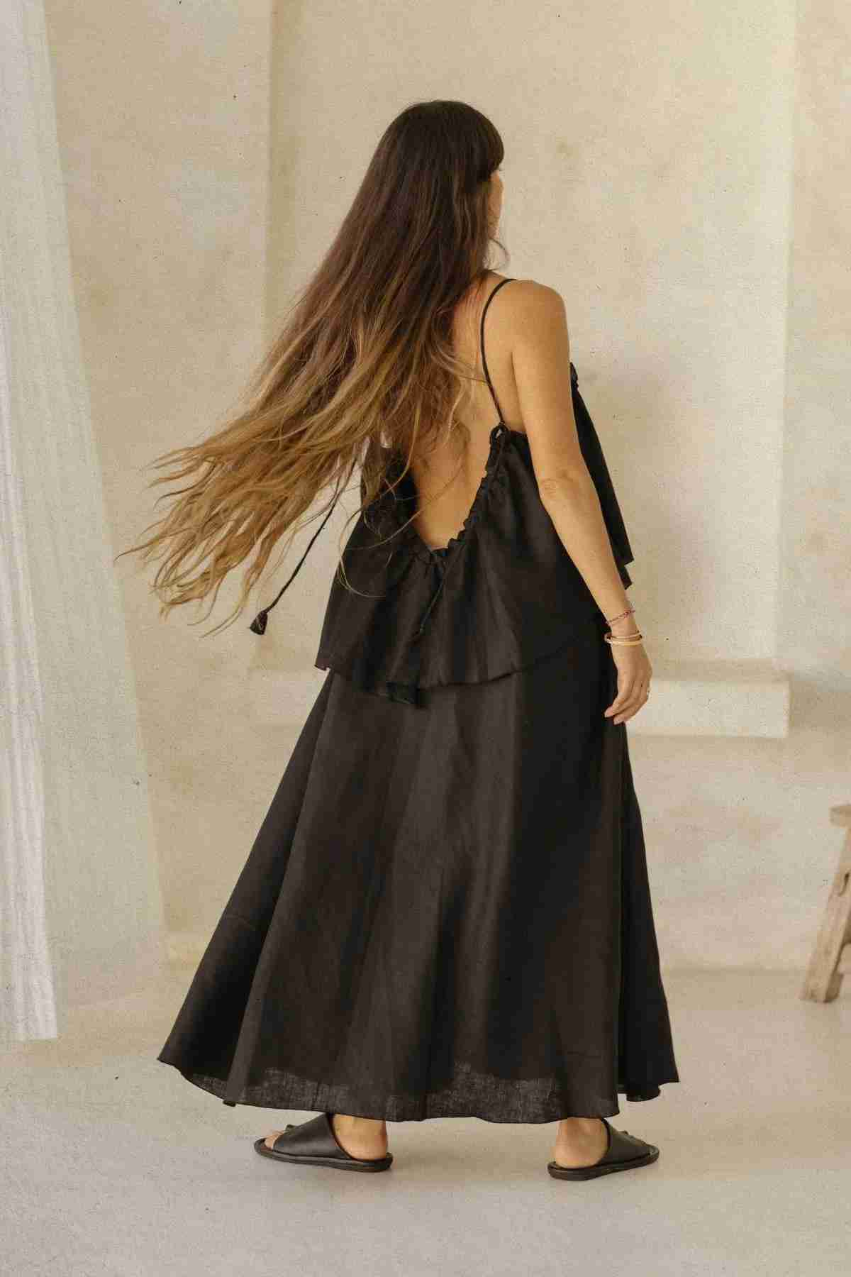 A woman with long brown hair, seen from behind, is sporting the Linen Muse Gown by Myrah Penaloza. The flowing dress features adjustable straps and an open back, enhancing its layered design. She stands barefoot indoors, facing a light-colored wall, looking relaxed in her stone-washed linen attire.