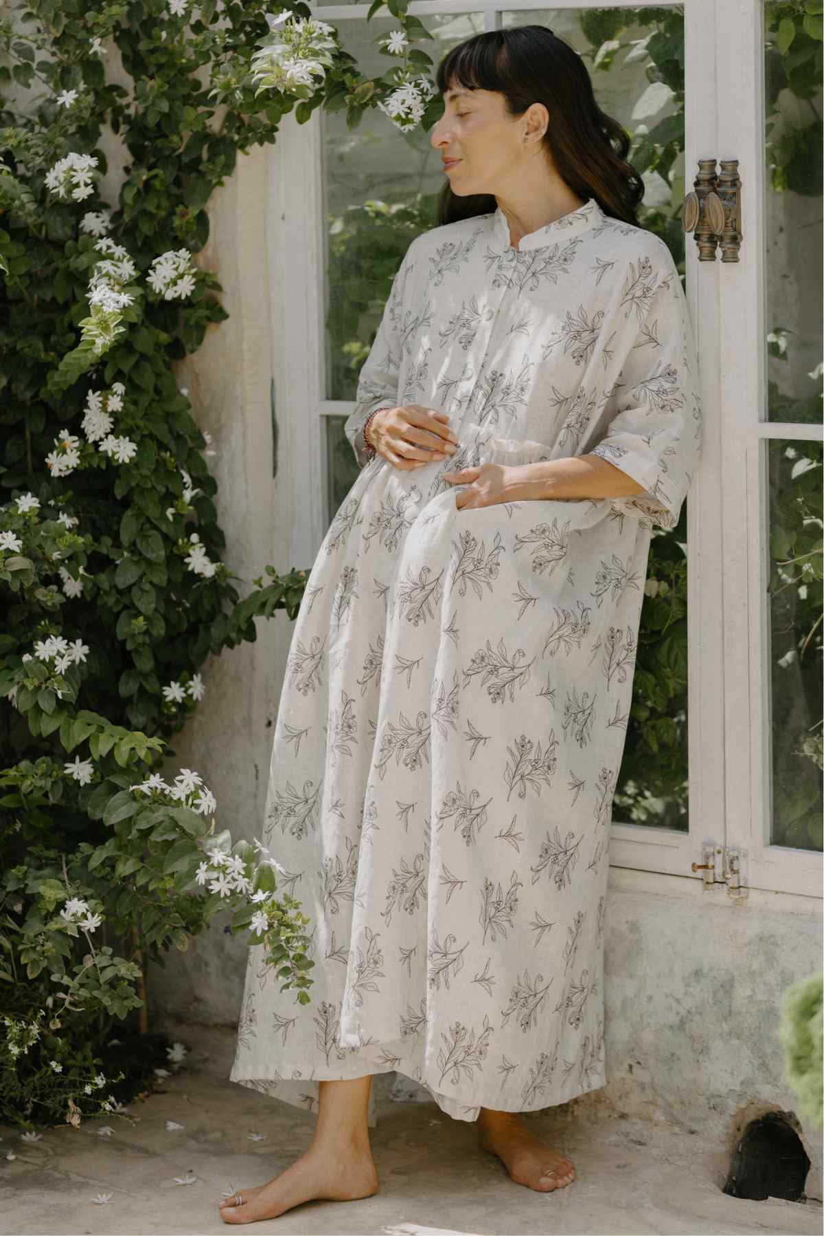 A pregnant woman stands barefoot outside, wearing the flowing, long-sleeved Bona Dress from the Cha Dao Tea Collection by Myrah Penaloza, adorned with a delicate floral pattern. She gracefully touches her belly, surrounded by lush green foliage and white flowers that contribute to the serene and natural ambiance.