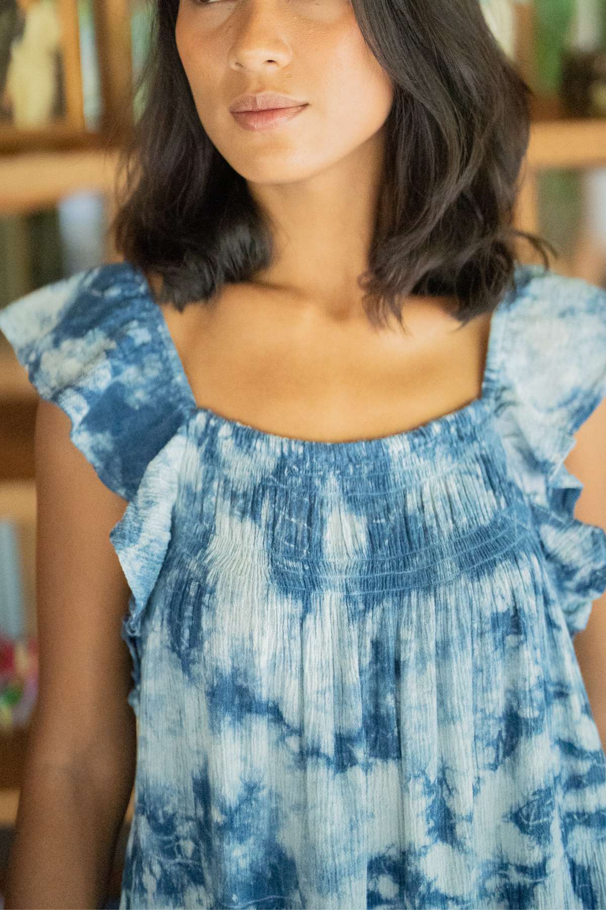 A woman with shoulder-length dark hair is seen wearing the Indigo Dreams Anna Yemaya Gown by Myrah Penaloza, a blue and white tie-dye piece made from crinkle cotton and featuring ruffled sleeves. In the background, blurry bookshelves filled with various books add to the scene.