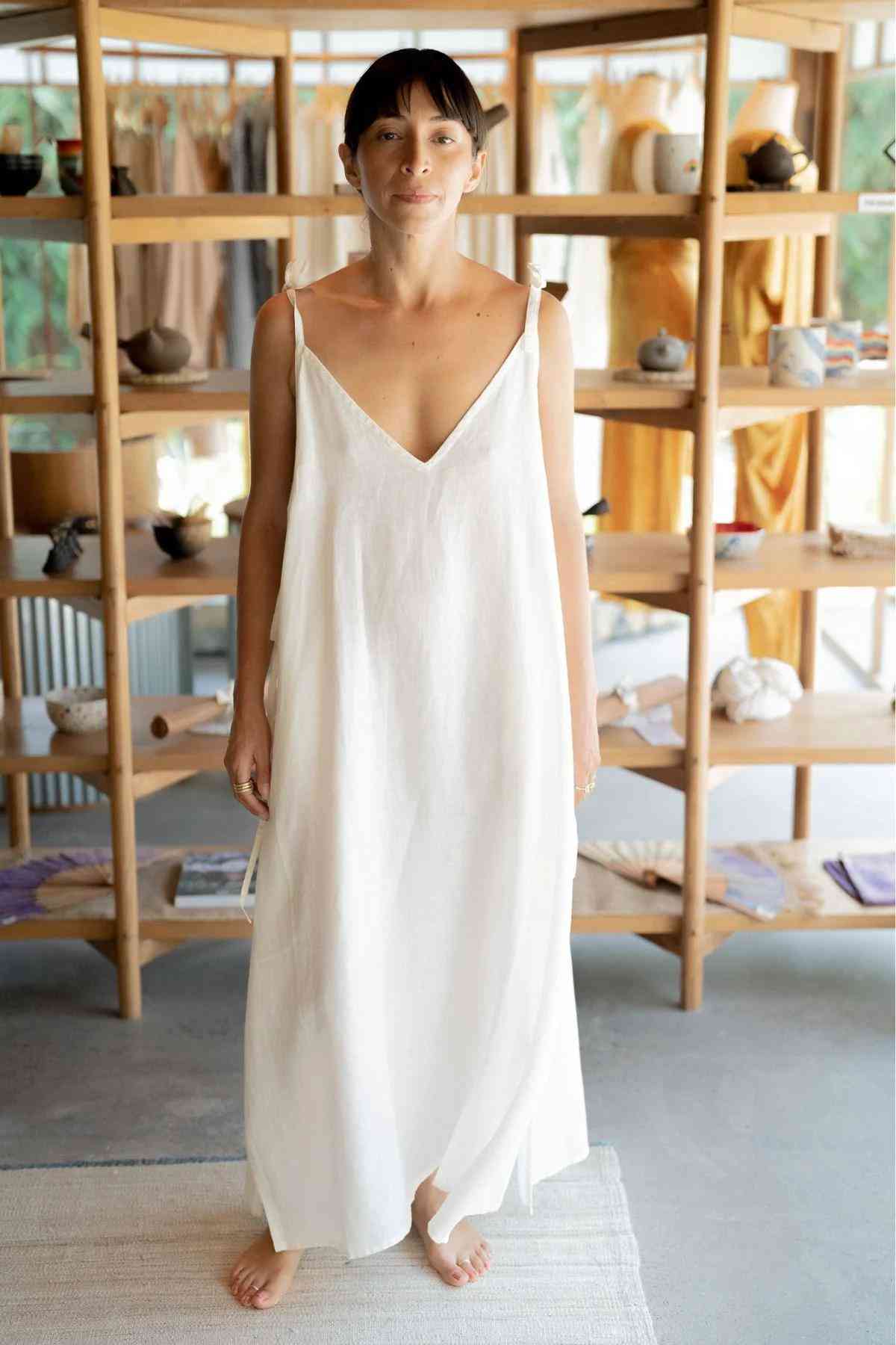 A person stands barefoot in a room with wooden shelves, wearing the Radiant Gown from the Myrah Penaloza healing linen collection. The long, sleeveless white dress with a V-neckline complements the backdrop of fabric, books, and pottery displayed on the shelves. The room has a light and airy ambiance.