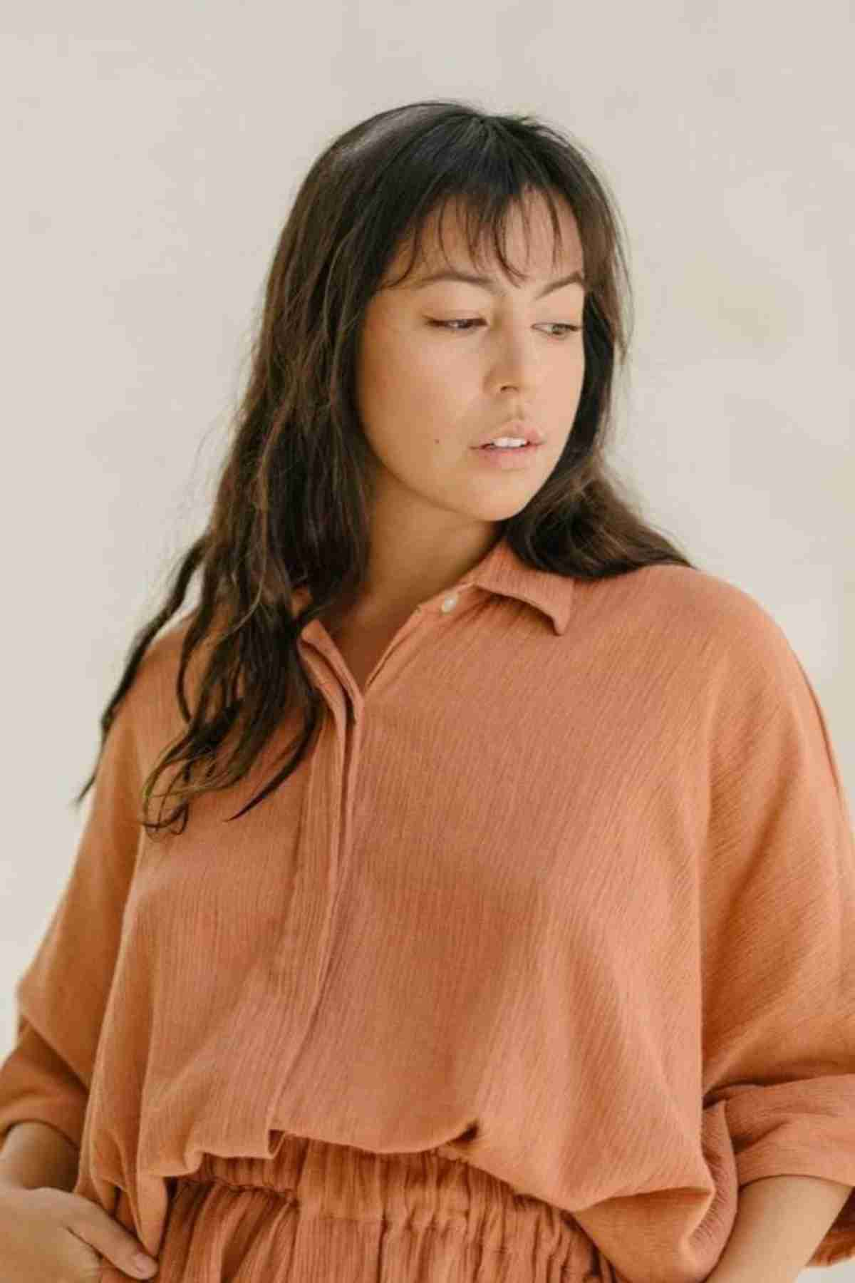 A person with long dark hair is gazing sideways while wearing the Suka Button Down Top from Myrah Penaloza, a loose-fitting blouse in a rust color with folded sleeves. This piece, handmade in Bali using plant-dyed methods, stands out against the soft, neutral-toned background.