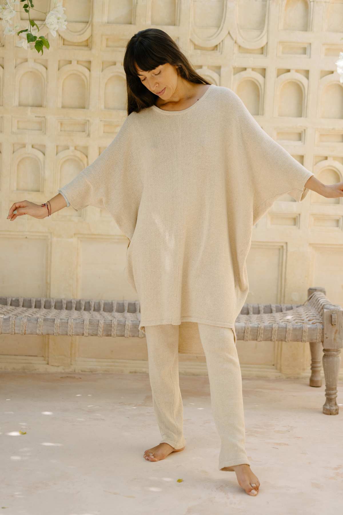 A woman stands posed with her arms slightly extended, wearing a loose, beige Butterfly Tunic Cotton Knit top by Myrah Penaloza and matching pants. She is in front of a light-colored, ornate wall with arches and a bench. The scene exudes a soft, serene ambiance.