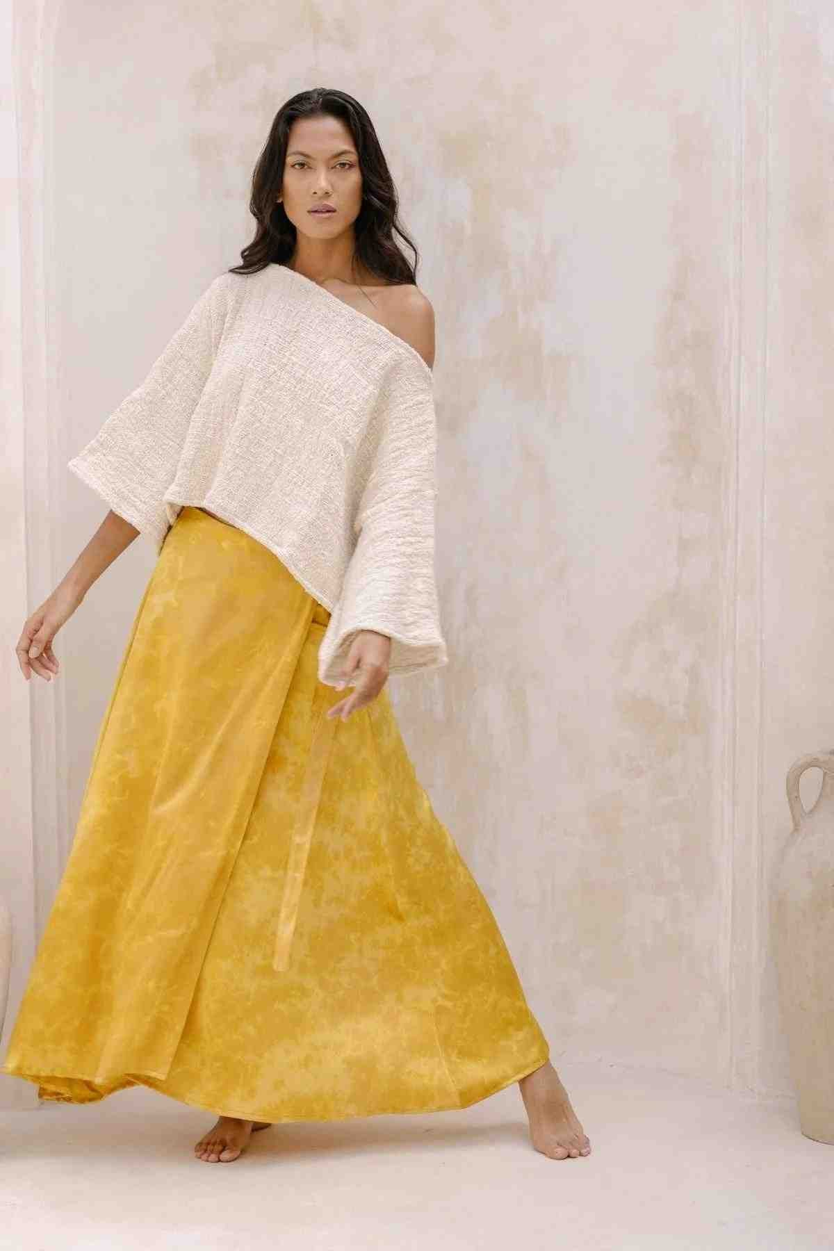 A woman poses in front of a neutral-toned background wearing the Spirit Top & Shanti Wrap Skirt Set by Myrah Penaloza. The off-the-shoulder, hand-woven Tummangal cotton top pairs beautifully with the flowing, ankle-length adjustable yellow skirt, creating a relaxed and elegant style. She stands barefoot, striking a confident pose.