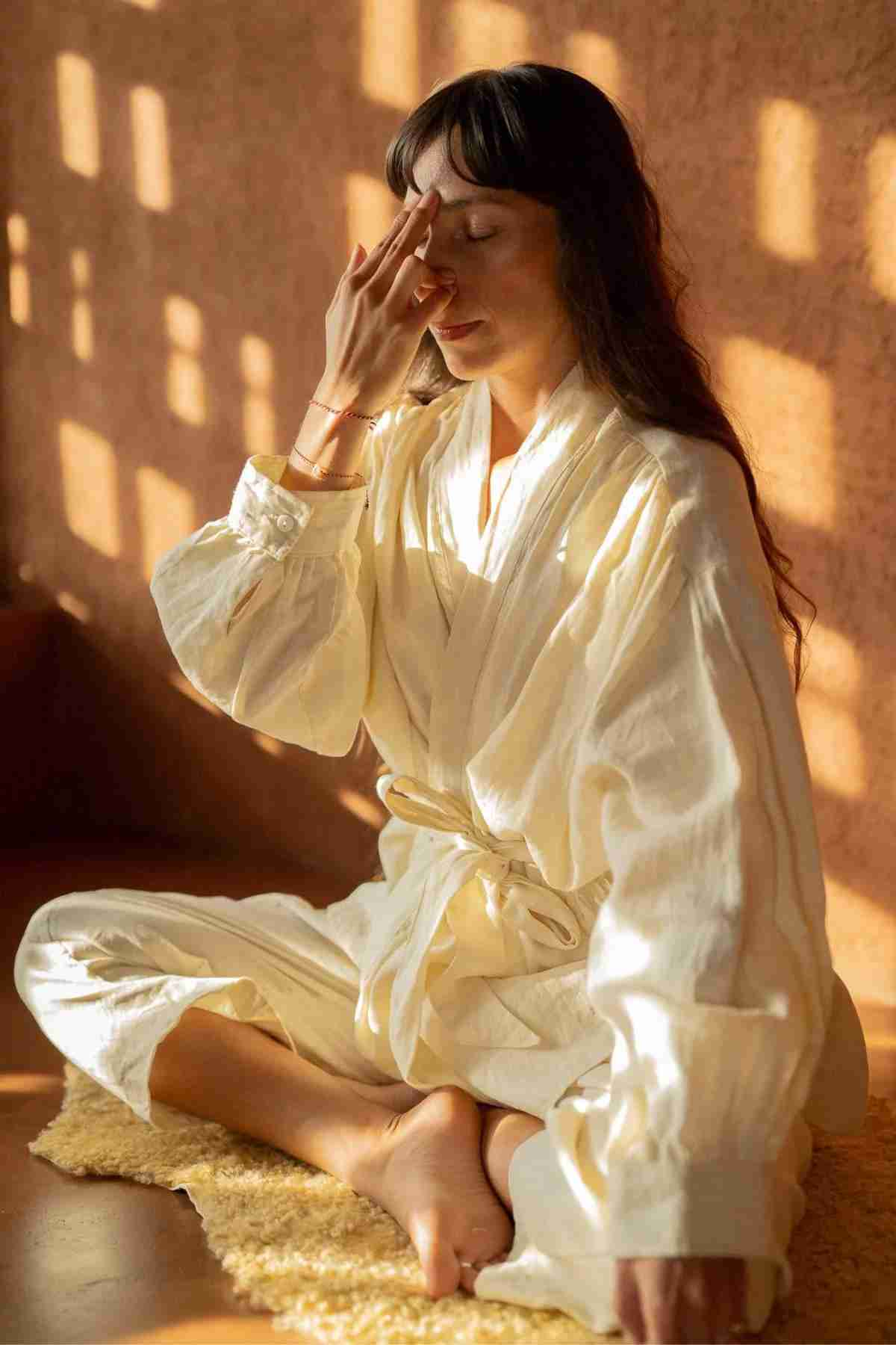 A woman with long hair sits cross-legged on a textured mat, wearing the Linen Jasmine Set by Myrah Penaloza. She is indoors, gently touching one side of her nose with closed eyes and a calm expression, as sunlight filters through a patterned window, casting shadows on the wall.