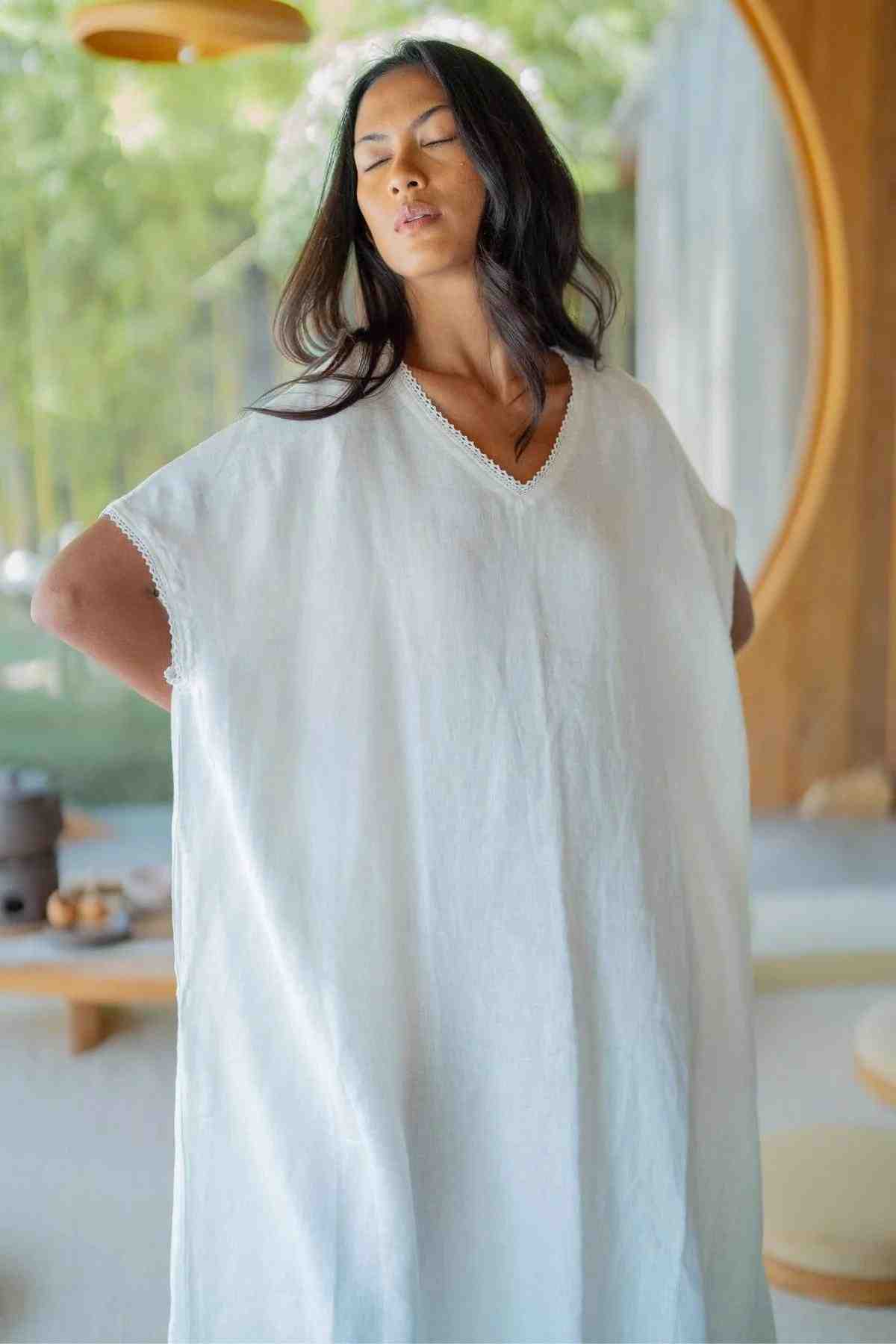 A person with long, dark hair closes their eyes while posing indoors. They are wearing the Ruby Lace Tunic by Myrah Penaloza, featuring a loose fit and a light-colored lace fabric with a V neckline. In the blurred background, wooden furniture is visible alongside greenery seen through a large window, creating a calm and serene ambiance.