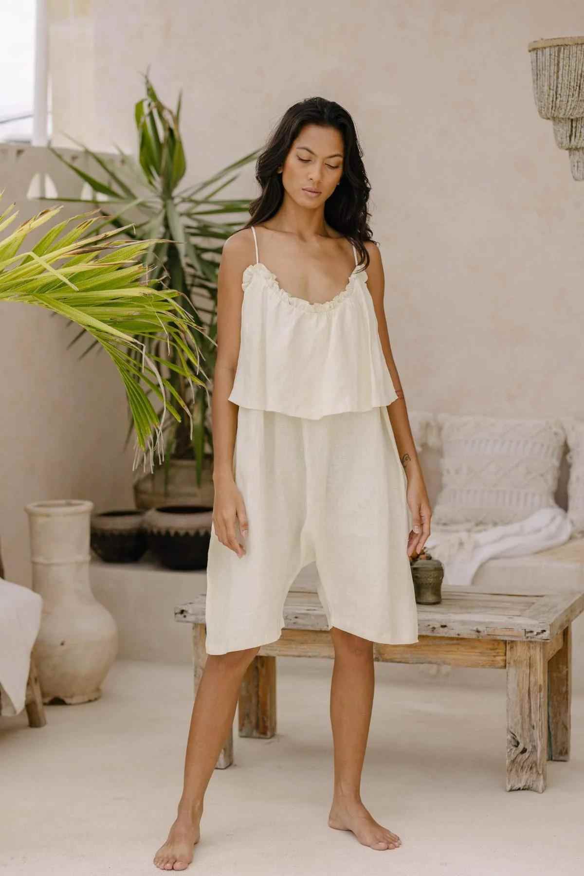 A woman stands barefoot in a relaxed, airy room adorned with plants and neutral-toned decor. She is wearing the Muse Linen Playsuit (Pre-Order) by Myrah Penaloza, featuring a light, loose-fitting cream-colored design with adjustable straps and a low back cut. Behind her is a wooden table with a decorative pillow, and to the left, a large green plant.