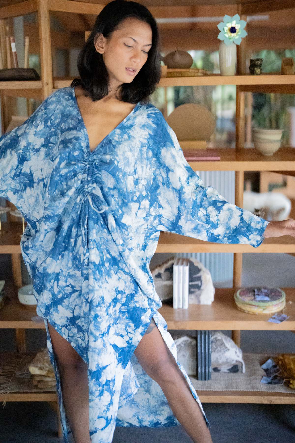 Draped in the Indigo Virgo Rising Kaftan (One of a Kind) by Myrah Penaloza, a person stands elegantly in front of a wooden shelf adorned with various decorative items. The kaftan's blue and white tie-dye patterns exude a seamless blend of elegance and style as they extend one arm in a poised gesture.