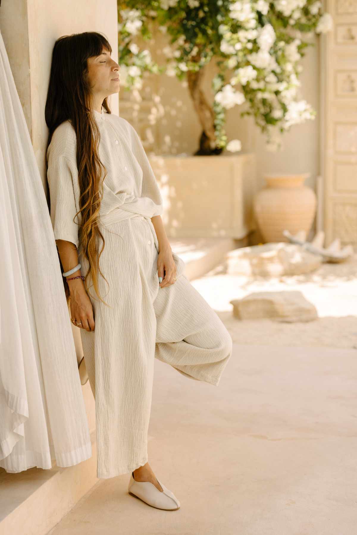 A woman with long brown hair stands serenely against a light-colored wall, eyes closed, wearing the Pine Playsuit by Myrah Penaloza. She is in a sunlit, peaceful setting with plants and neutral-toned decor that complements her versatile wardrobe.