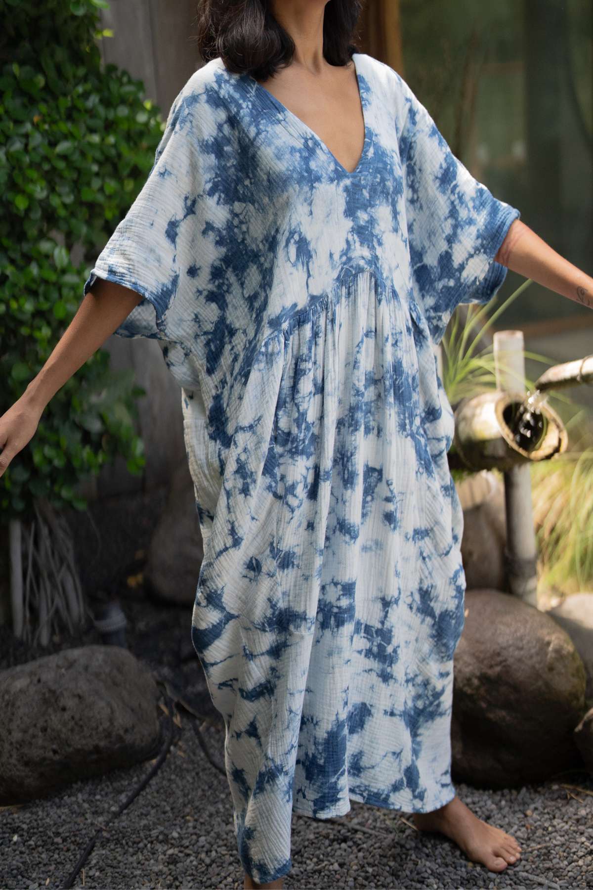 Wearing the Indigo Venus Gown by Myrah Penaloza, a person stands on gravel outdoors. The gown features a blue and white tie-dye pattern in cotton gauze, with a V-neck and loose sleeves. Embracing an effortless style, they have their arms outstretched amid lush greenery and rocks.