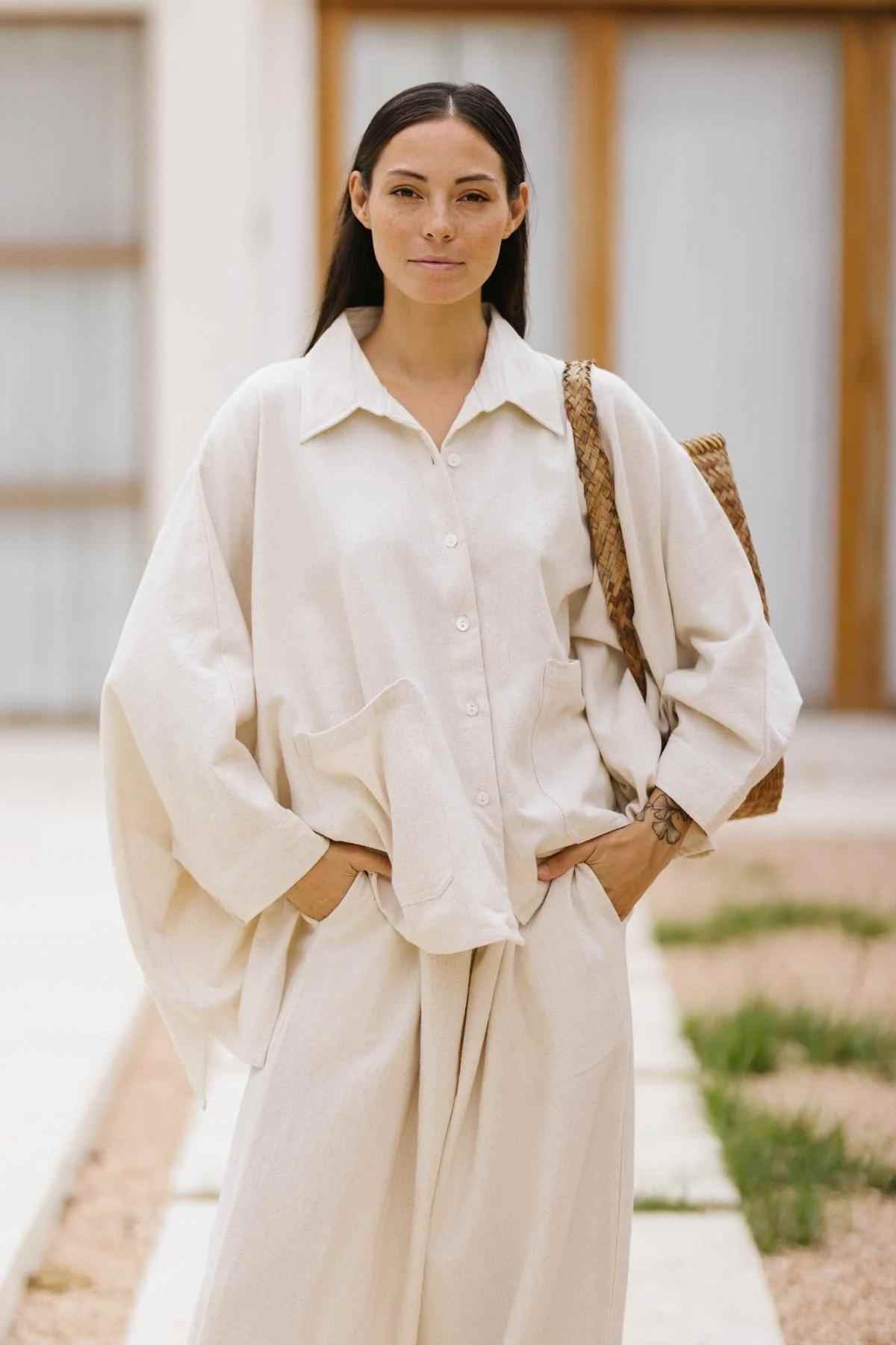 A person with long, dark hair stands in an outdoor setting. They are wearing a loose, 100% cotton linen Grace Jacket and Mona Pant Set in a natural beige hue from Myrah Penaloza, made to order. Their hands rest in their pockets as they carry a woven bag over one shoulder. In the background, there is a building and a garden path.