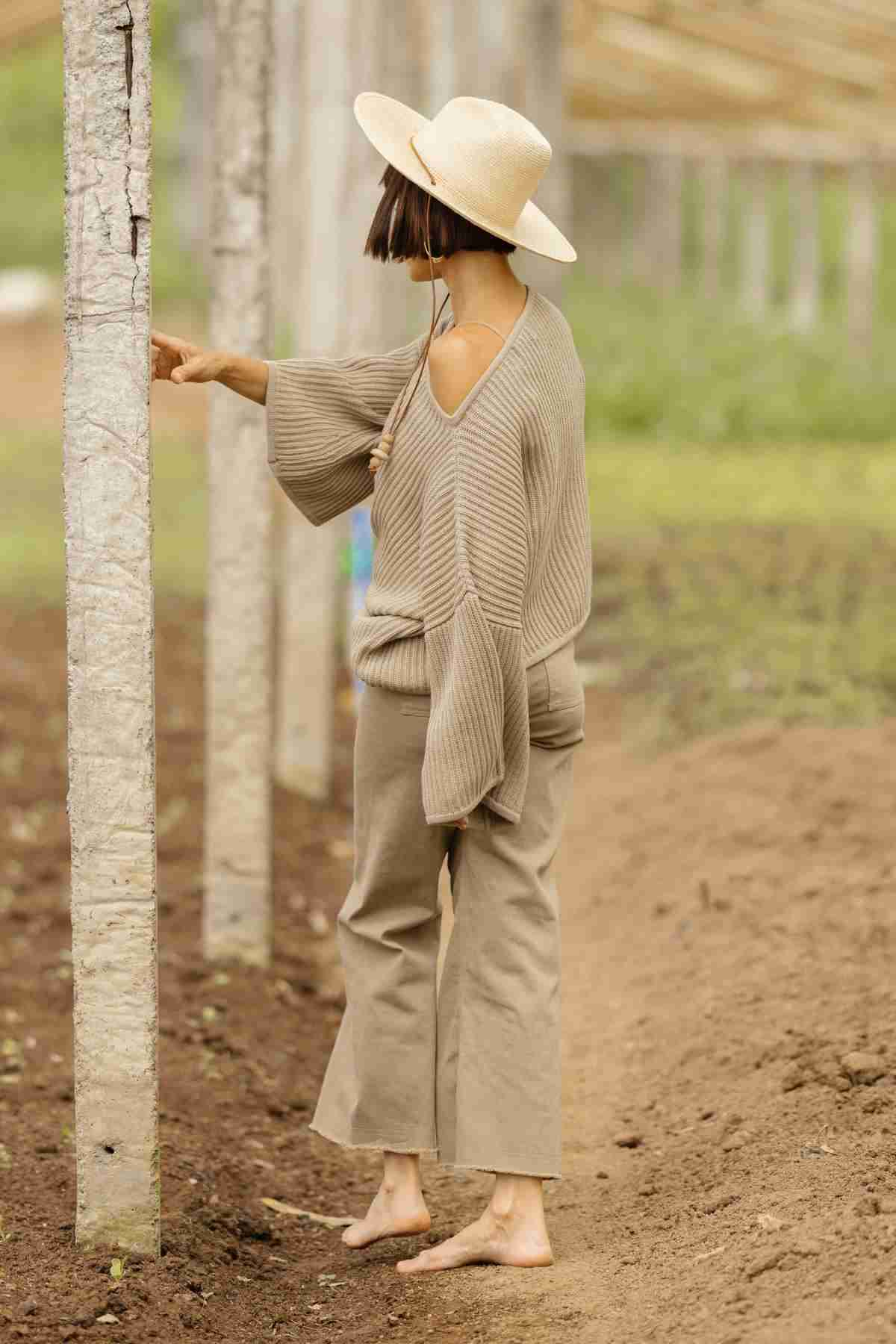 A person in a beige wide-brimmed hat, the Spirit Top Sweater Knit by Myrah Penaloza, and loose beige pants stands barefoot on a dirt path. They are touching a concrete post with one hand, while their head is turned slightly to the side, looking at the post. The background is green and brown.