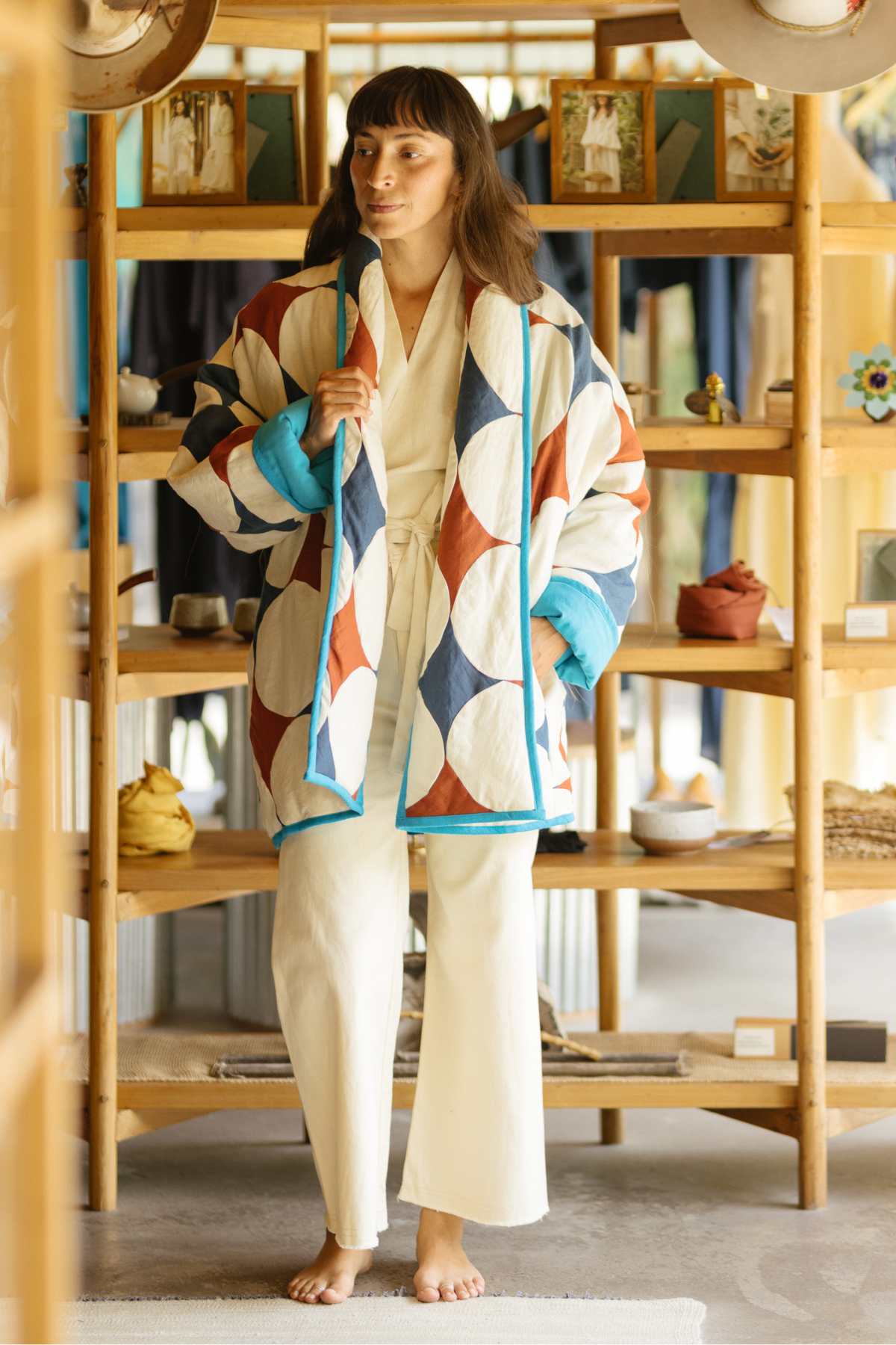 A person stands in front of wooden shelves wearing a Myrah Penaloza Javi Quilted Jacket (Pre-Order) featuring large geometric patterns in blue, white, and brown. They are also dressed in light-colored, possibly white, casual clothing underneath. The background shows various neatly displayed items.