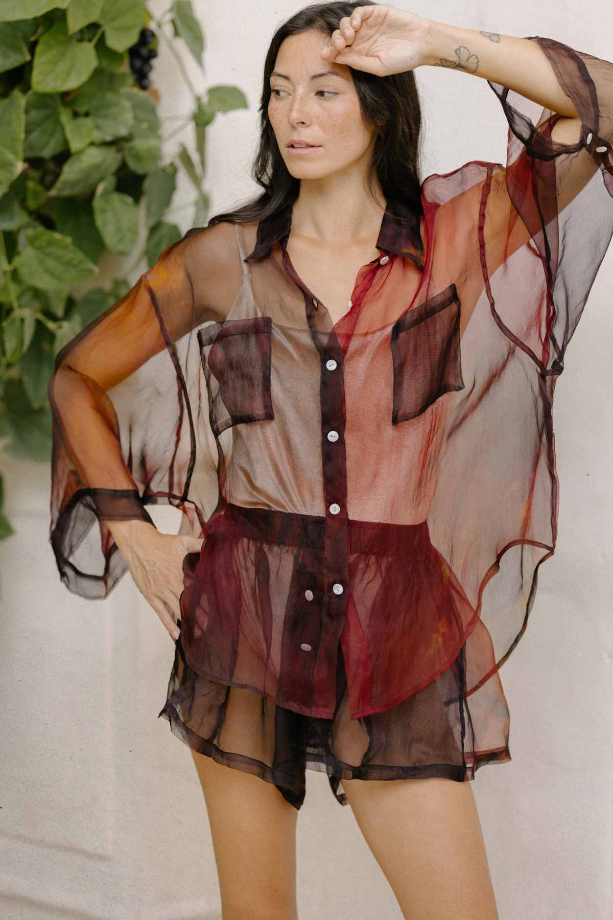 A person poses in the Rainbeau Nidra Chiffon Set by Myrah Penaloza, featuring a sheer, flowy, long-sleeve blouse with a dark collar and buttons, paired with matching sheer shorts. They have one hand on their head and stand against a backdrop of green foliage and a light-colored wall.