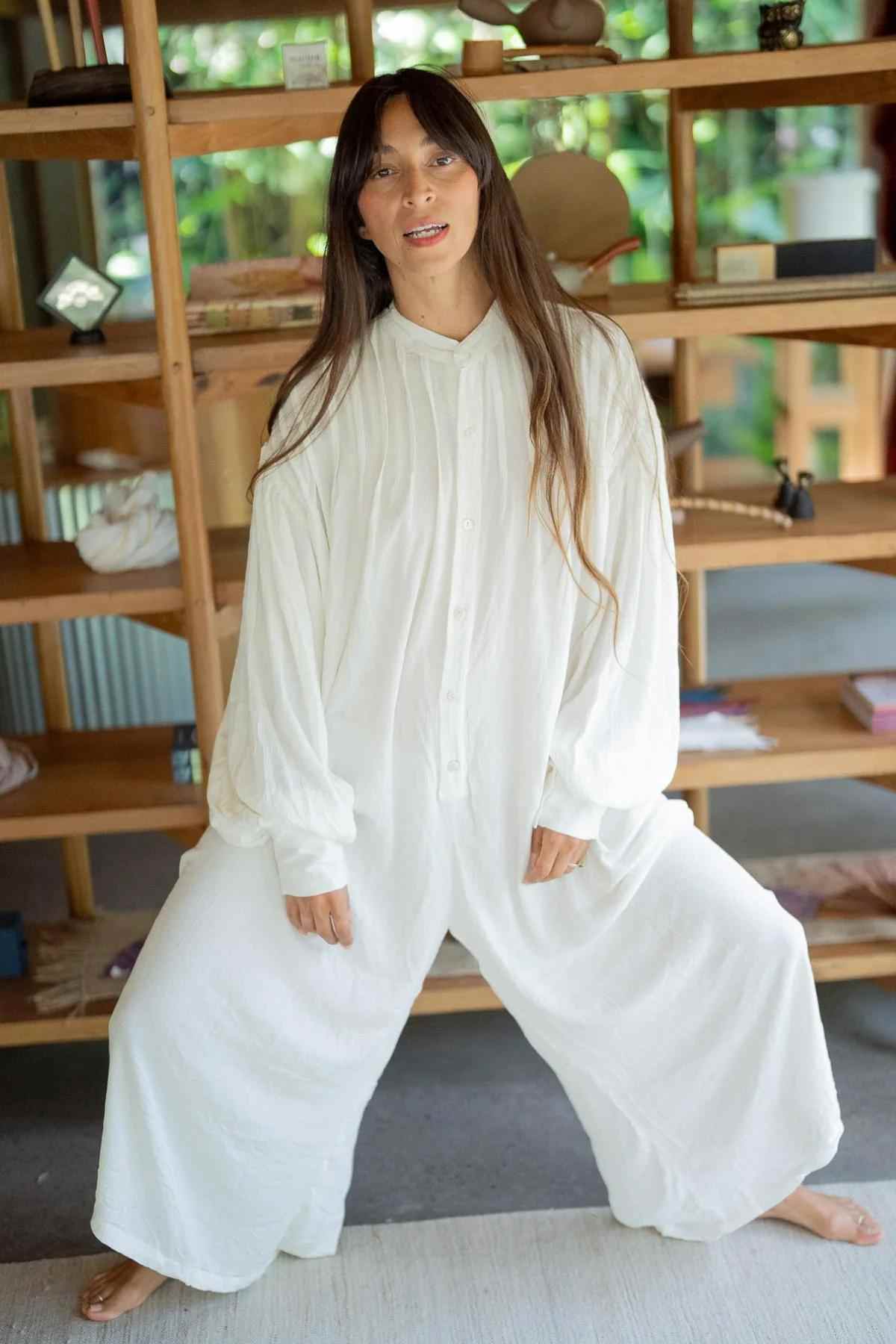 A person with long hair is standing in a relaxed pose, wearing a loose-fitting Kundalini Gown Playsuit Edition (Pre-Order) by Myrah Penaloza, which features wide legs and long sleeves. They are standing in front of a wooden bookshelf filled with various objects. The background shows a brightly lit, cozy indoor space.