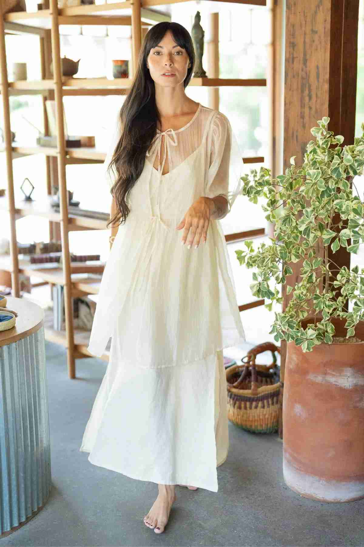 A barefoot woman with long dark hair wears a Myrah Penaloza Dynasty Sheer Silk Dress, a flowing white ensemble crafted from silk chiffon and featuring essential layering details with a bow at the neckline. She stands indoors in front of wooden shelves adorned with various decor and a tall potted plant, exuding a serene and natural vibe.