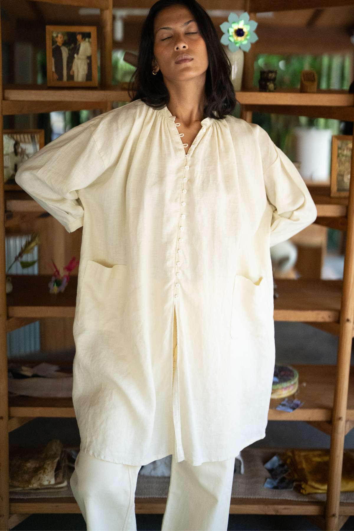 A person with long dark hair is wearing the Zazen Linen Top from Myrah Penaloza, exuding effortless elegance in its loose, cream-colored design over matching pants. They stand with their hands behind their back and eyes closed, set against a background of wooden shelves adorned with various small items.