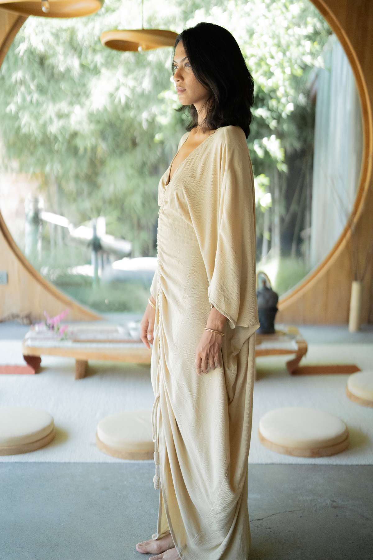 A woman in an elegant kaftan by Myrah Penaloza, named Virgo Moon, stands indoors near a large circular window. Sunlight filters through, highlighting bamboo and a serene garden outside. Inside, round cushions and a small wooden table with a teapot complete the tranquil scene.