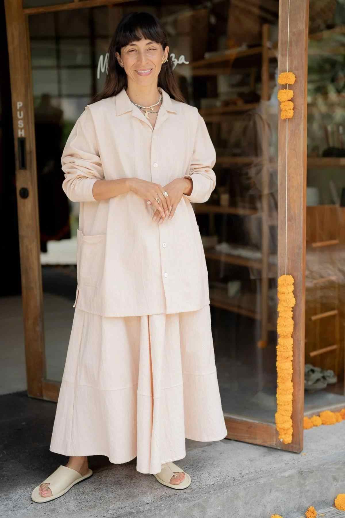 A person with long dark hair is standing in front of a wooden door adorned with orange marigold flowers. They are wearing the Tender Jacket & Pant Set (3 of a kind) - Pre Order Only by Myrah Penaloza, featuring a blush pink jacket paired with slide sandals. The background includes wooden shelves inside the building.