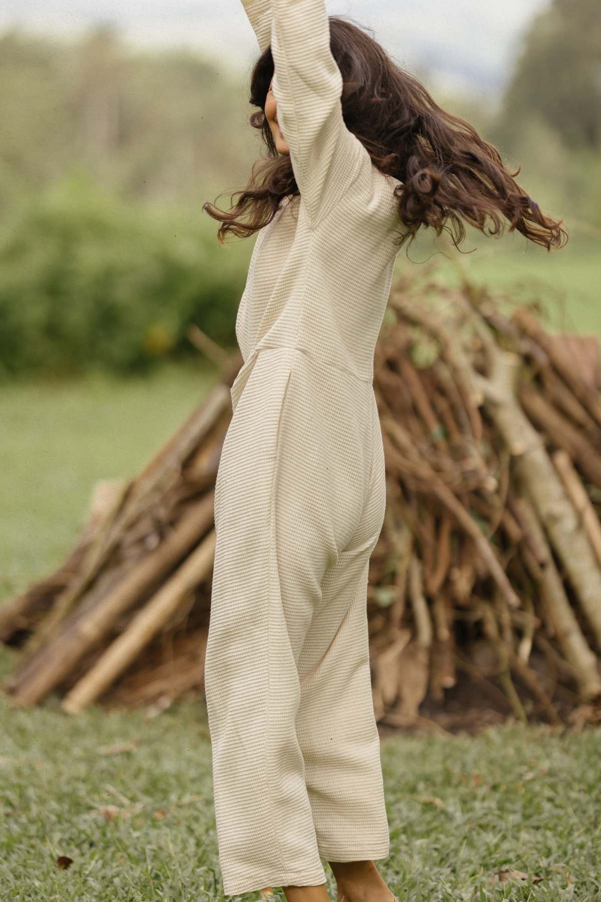 A person with long, wavy hair wearing the Myrah Penaloza Camper Playsuit is captured mid-jump on grassy ground. There's a pile of logs in the background amidst a lush, green outdoor setting—perfect for adventurous weekend getaways.