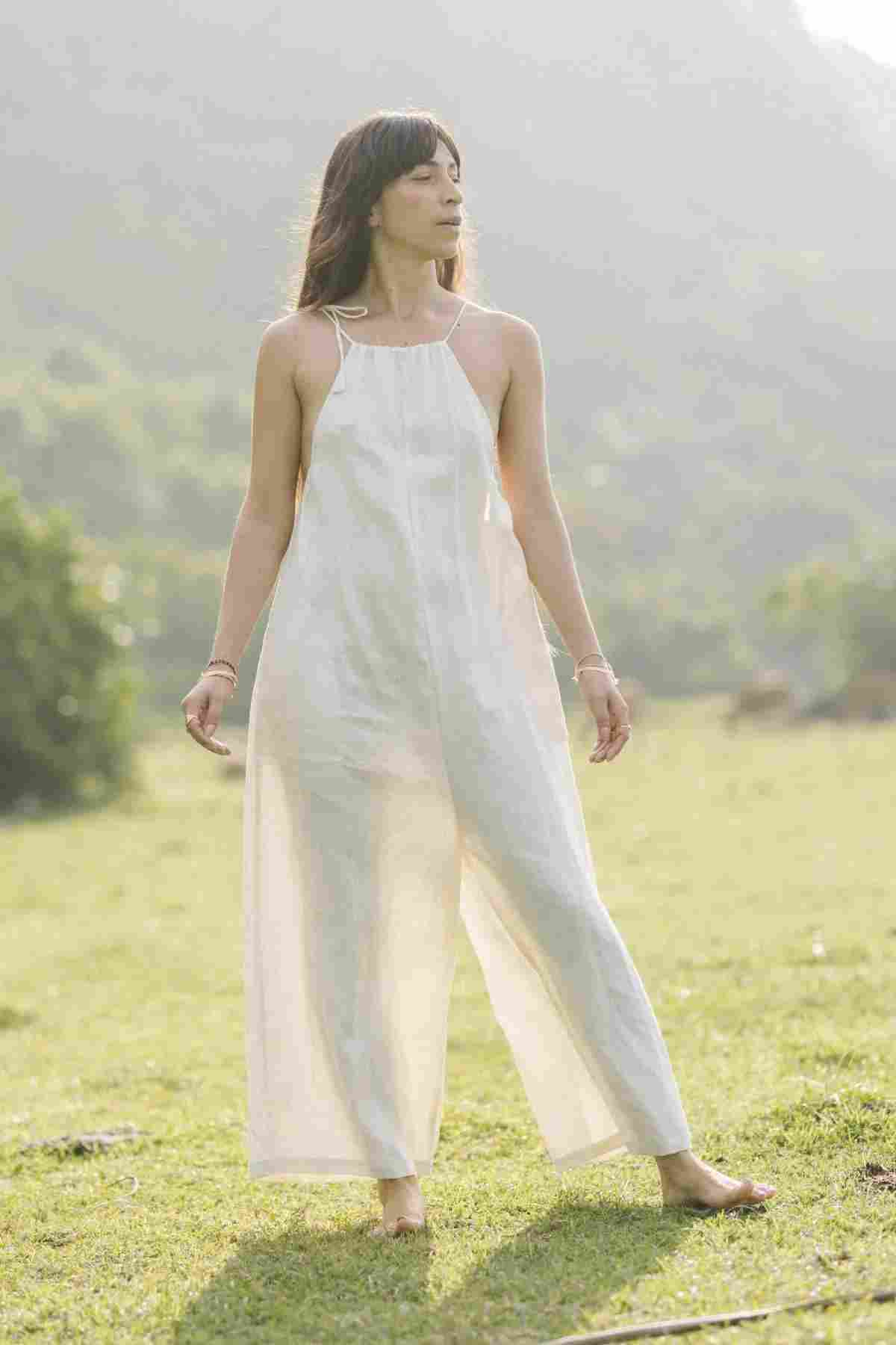 A person with long hair stands outside in a grassy field with trees and hills in the background. They are wearing a flowing, sleeveless, light-colored Linen Vata Playsuit by Myrah Penaloza made of 100% linen and are barefoot, gazing thoughtfully into the distance as sunlight filters through the scene.