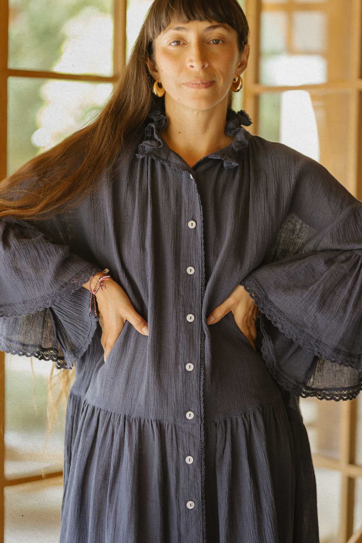 Indoors, a person with long hair poses confidently in The Juliette Linen Gown by Myrah Penaloza. This elegant garment, crafted from a blend of linen and cotton, is adorned with ruffled sleeves and comes in a captivating blue hue. With hands on their hips, they gaze directly at the camera as large windows behind them illuminate the space with soft, warm light.