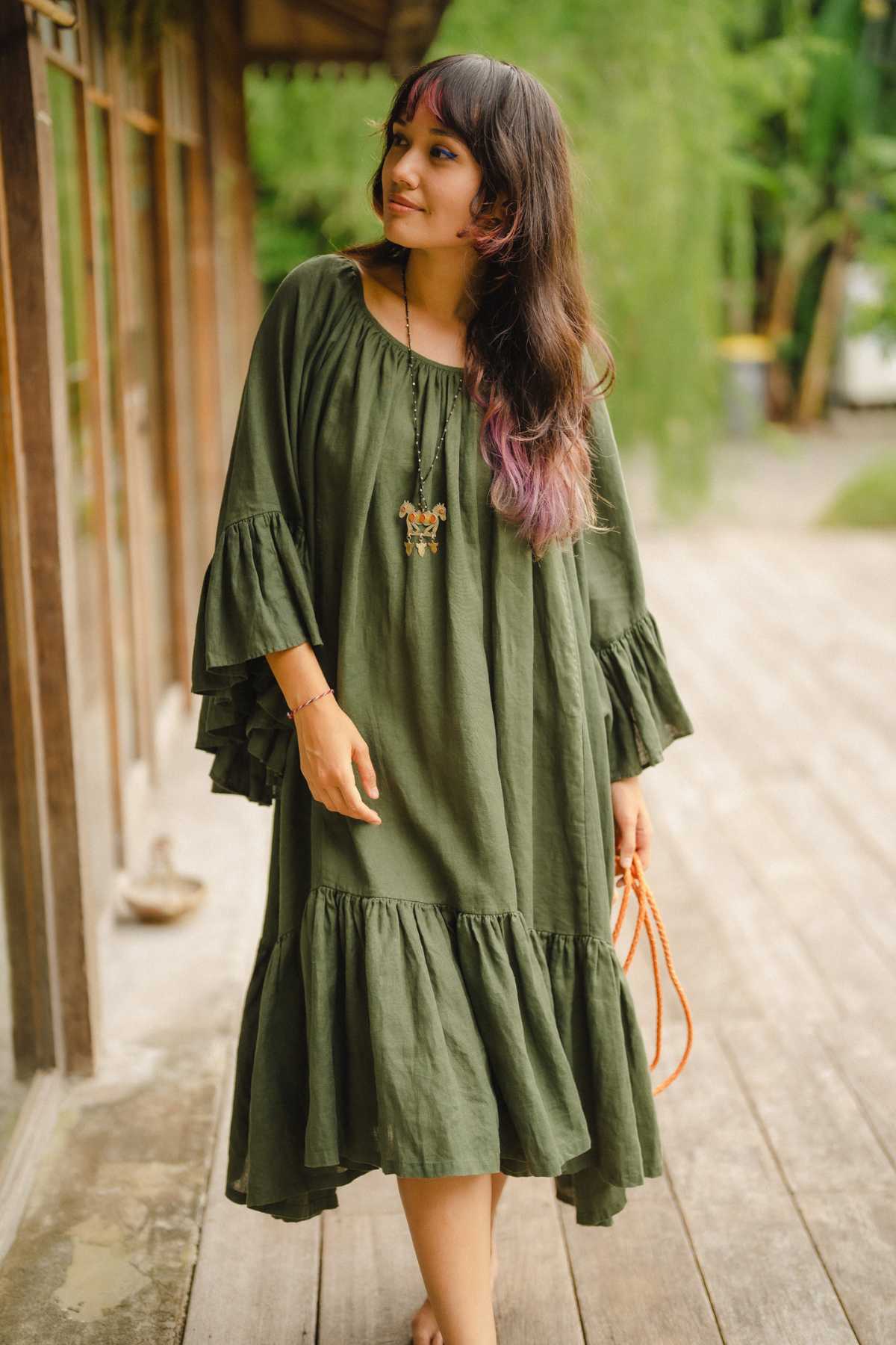 A woman in a flowing Brida Linen Gown Long by Myrah Penaloza gracefully strolls along a wooden path beside a building. Her long hair with purple highlights complements her pendant necklace, perfectly framed by the lush greenery surrounding her enchanting presence.