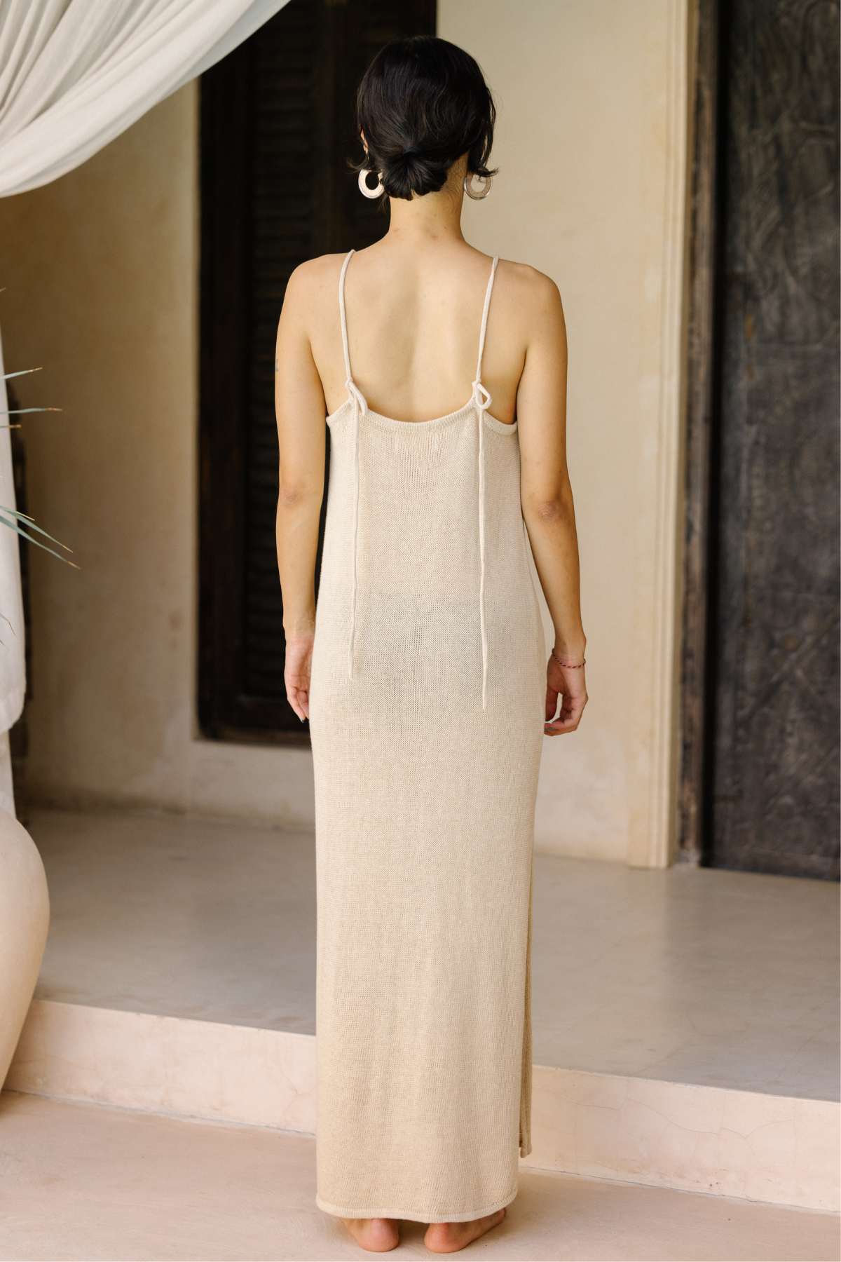 A person stands with their back facing the camera, wearing the Virgo Slip Hand Knit Cotton from Myrah Penaloza. The long, beige, sleeveless dress made of soft cotton knit features thin straps and a low back. The scene is set indoors with neutral-toned walls and a large ceramic vase visible on the left side of the image.