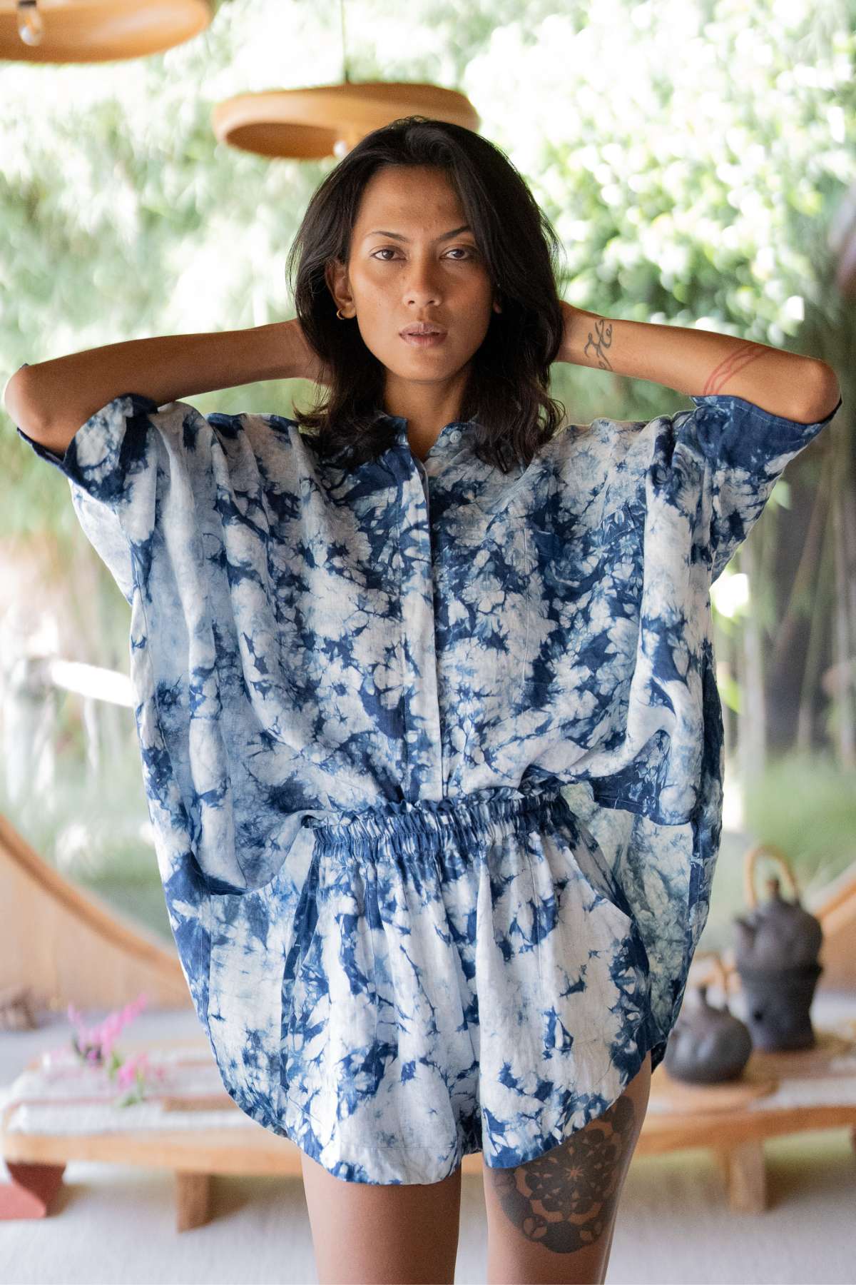 A person poses indoors, effortlessly chic in the Indigo Suka Set by Myrah Penaloza, featuring a blue and white tie-dye button-up shirt and matching shorts made from 100% linen sourced ethically. With hands behind their head, they stand against a backdrop of green foliage and modern decor elements.